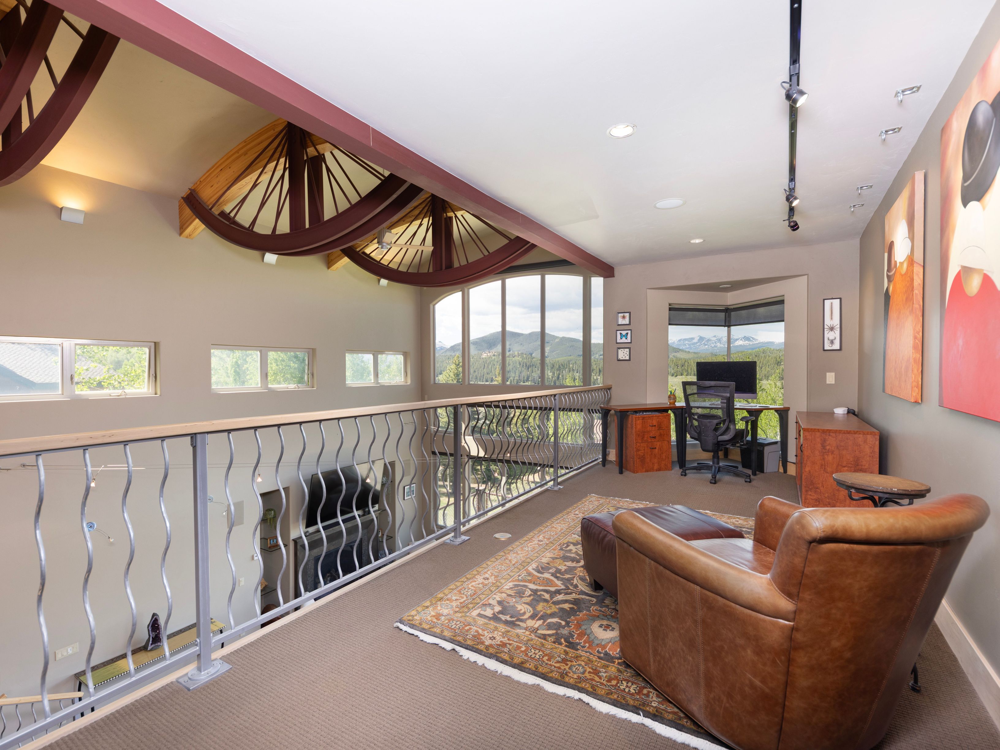 This spacious loft area serves as a home office, featuring a desk positioned against a wall of windows that frame expansive mountain views. The room is accented by unique curved structural beams overhead and a comfortable leather armchair with an ottoman resting on a patterned area rug. The open-concept design overlooks the lower level through a decorative metal railing, creating an airy and sophisticated workspace.