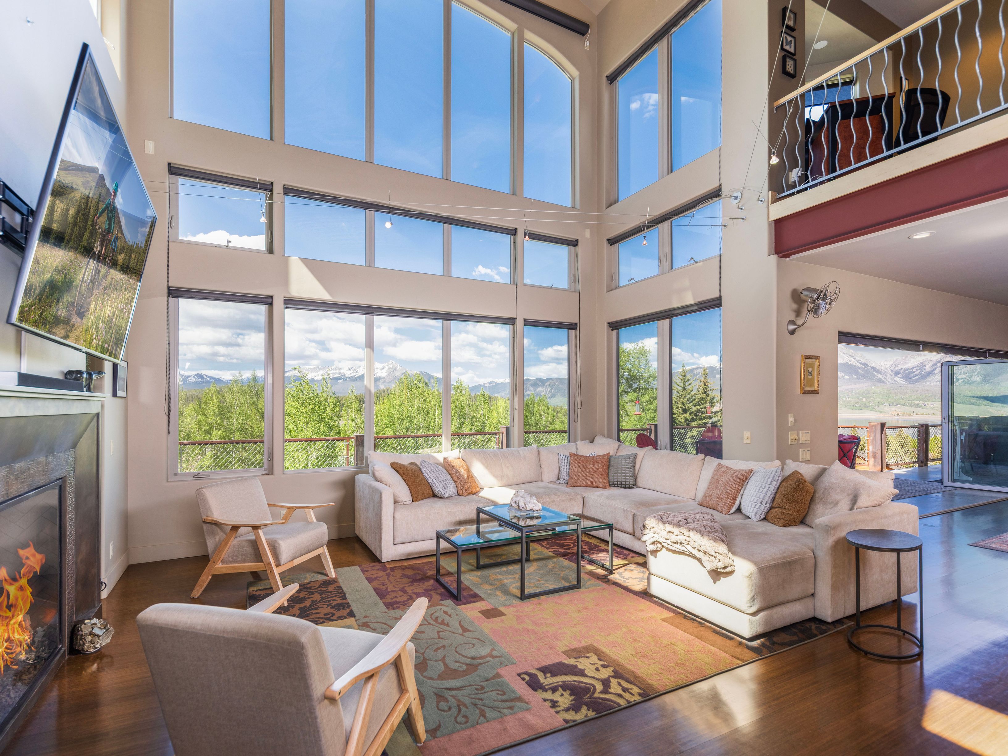 This expansive living room features a dramatic wall of tall windows that frame panoramic mountain views and flood the space with natural light. The room is anchored by a large, neutral-toned sectional sofa and a patterned area rug, complemented by two light wood armchairs and a modern fireplace with a dark surround. A mezzanine level with a metal railing overlooks the living area, enhancing the open, airy atmosphere of this mountain-style home.
