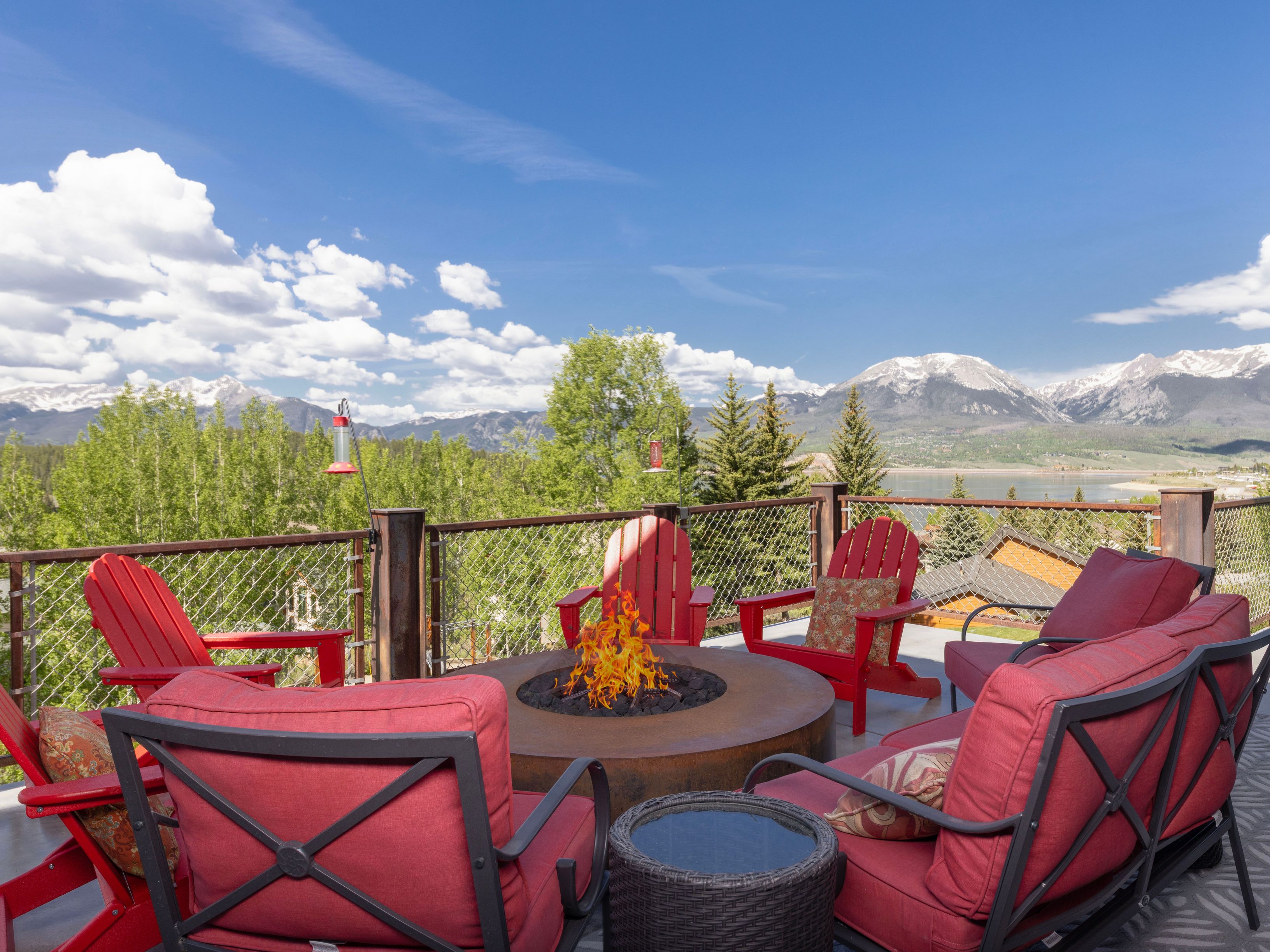 This elevated deck offers a stunning panoramic view of a lake and snow-capped mountains under a bright blue sky. The space is furnished with vibrant red Adirondack chairs and cushioned outdoor seating arranged around a circular, metal-rimmed fire pit. A cable railing with a wire mesh insert provides safety while maintaining the expansive, scenic vista.