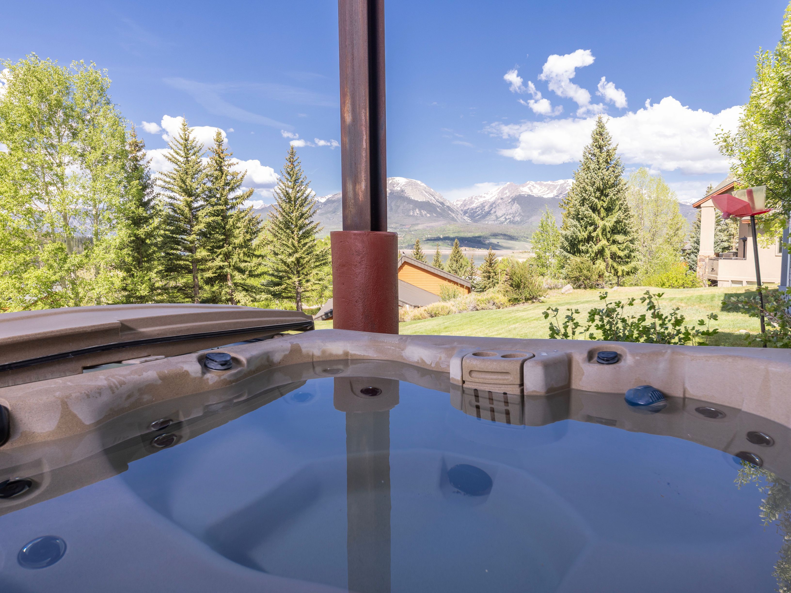 This image captures a relaxing hot tub situated on a deck, offering a stunning, elevated view of a mountain range and a distant lake. The foreground features the textured rim and water surface of the spa, while the background showcases a lush green landscape with evergreen trees and a clear blue sky. The perspective is from a seated position within the spa, emphasizing the tranquil and scenic outdoor living experience.