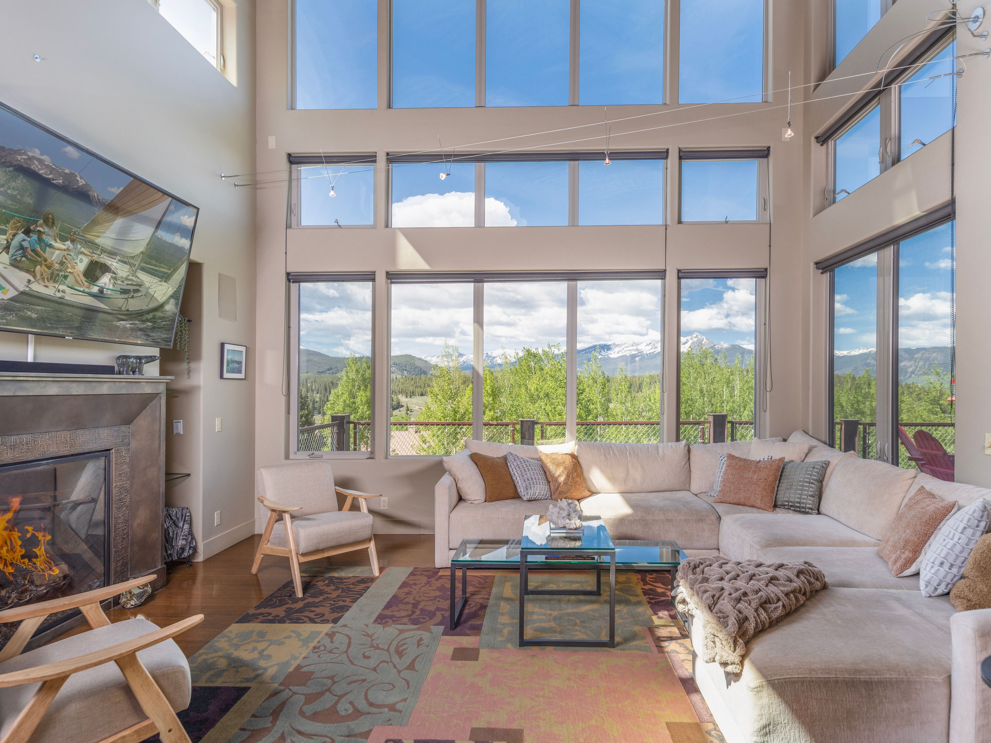 This spacious living room features a large sectional sofa and two light wood armchairs arranged around a glass-topped coffee table on a colorful patterned rug. A tall, metal-clad fireplace surround serves as a focal point, while a wall of expansive windows provides panoramic views of the surrounding mountains and forest. The room is bathed in natural light, creating an airy and inviting atmosphere perfect for relaxation.