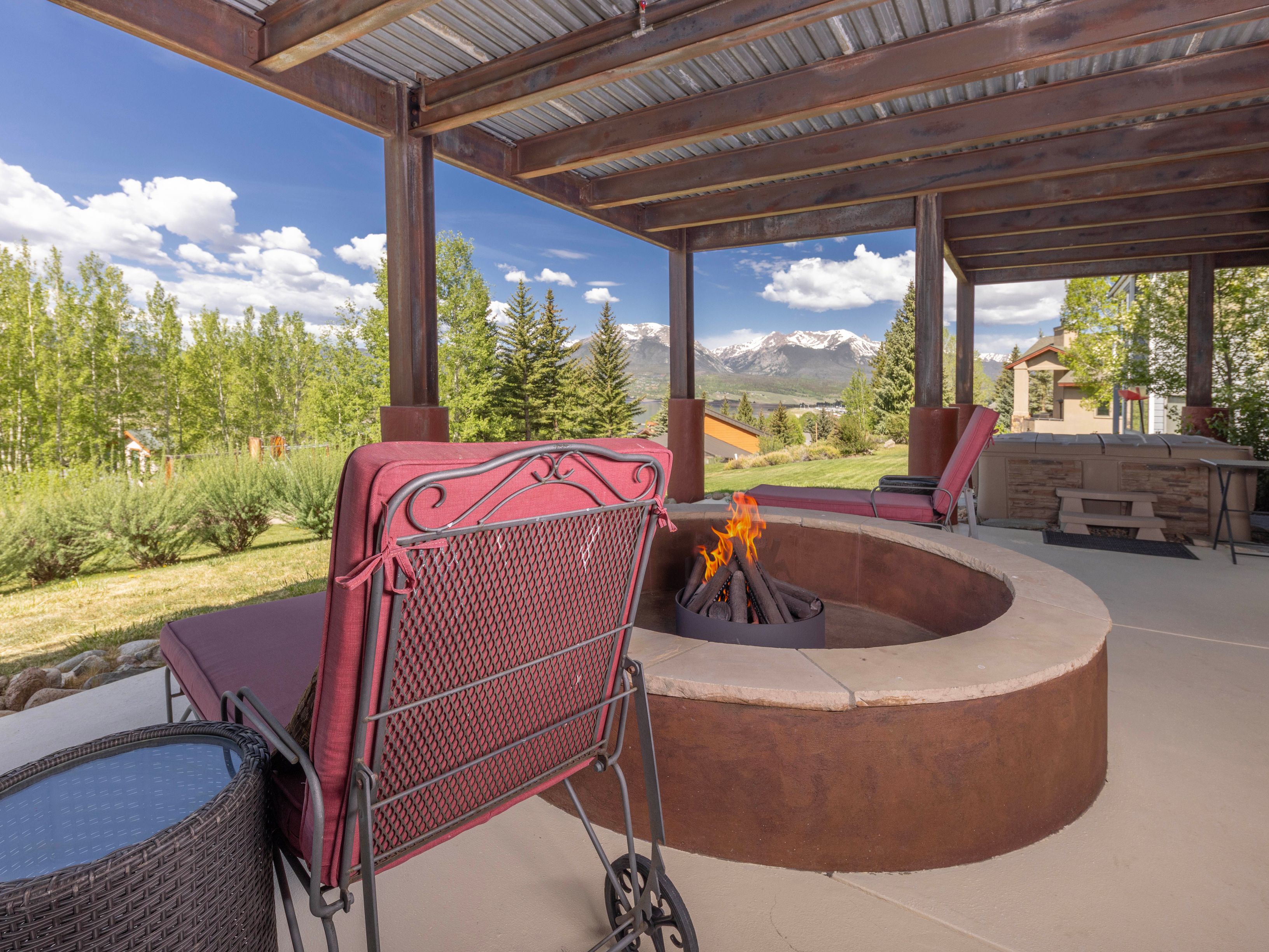 This covered patio offers a serene outdoor living space featuring a circular fire pit with a stone-topped rim and a rustic metal base. The area is furnished with comfortable lounge chairs and includes a hot tub tucked under the pergola structure, all set against a backdrop of lush green trees and snow-capped mountains. The open-air design provides a seamless connection to the surrounding landscape, creating an inviting atmosphere for relaxation.