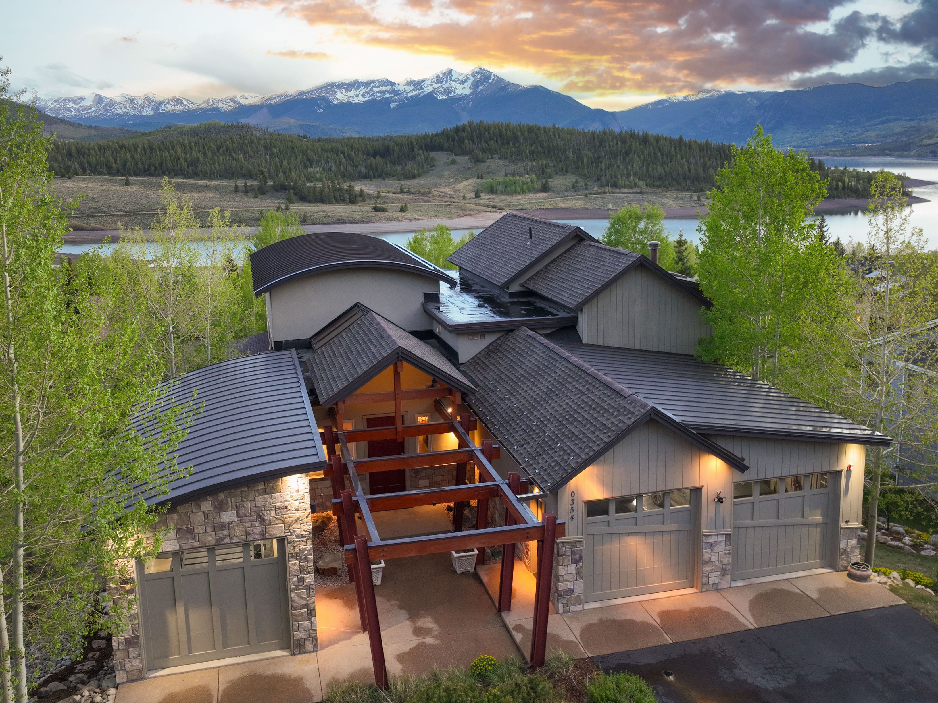 This elevated aerial perspective captures a modern mountain home set against a dramatic backdrop of snow-capped peaks and a serene lake at sunset. The residence features a complex roofline with a mix of metal and shingled surfaces, stone accents, and a prominent timber-framed entryway pergola. The warm glow of exterior lighting highlights the home's architectural details, creating an inviting and luxurious atmosphere in a picturesque natural setting.