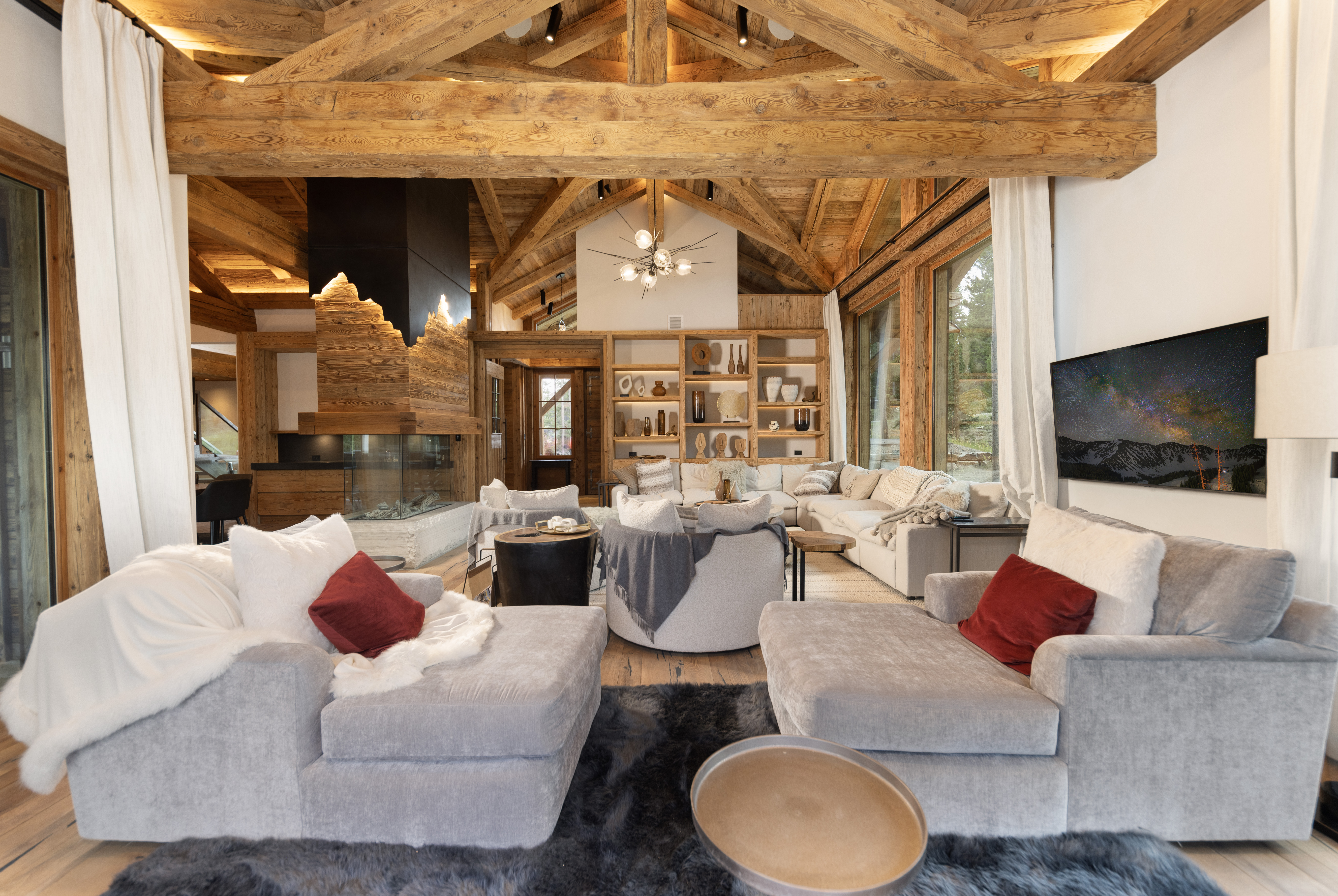 This is a spacious living room featuring a vaulted ceiling with exposed wooden beams, creating a rustic yet luxurious atmosphere. The room is furnished with comfortable gray sofas, a modern fireplace, and a built-in bookshelf, offering both style and functionality. Large windows provide ample natural light and views of the surrounding landscape, enhancing the overall appeal of the space.