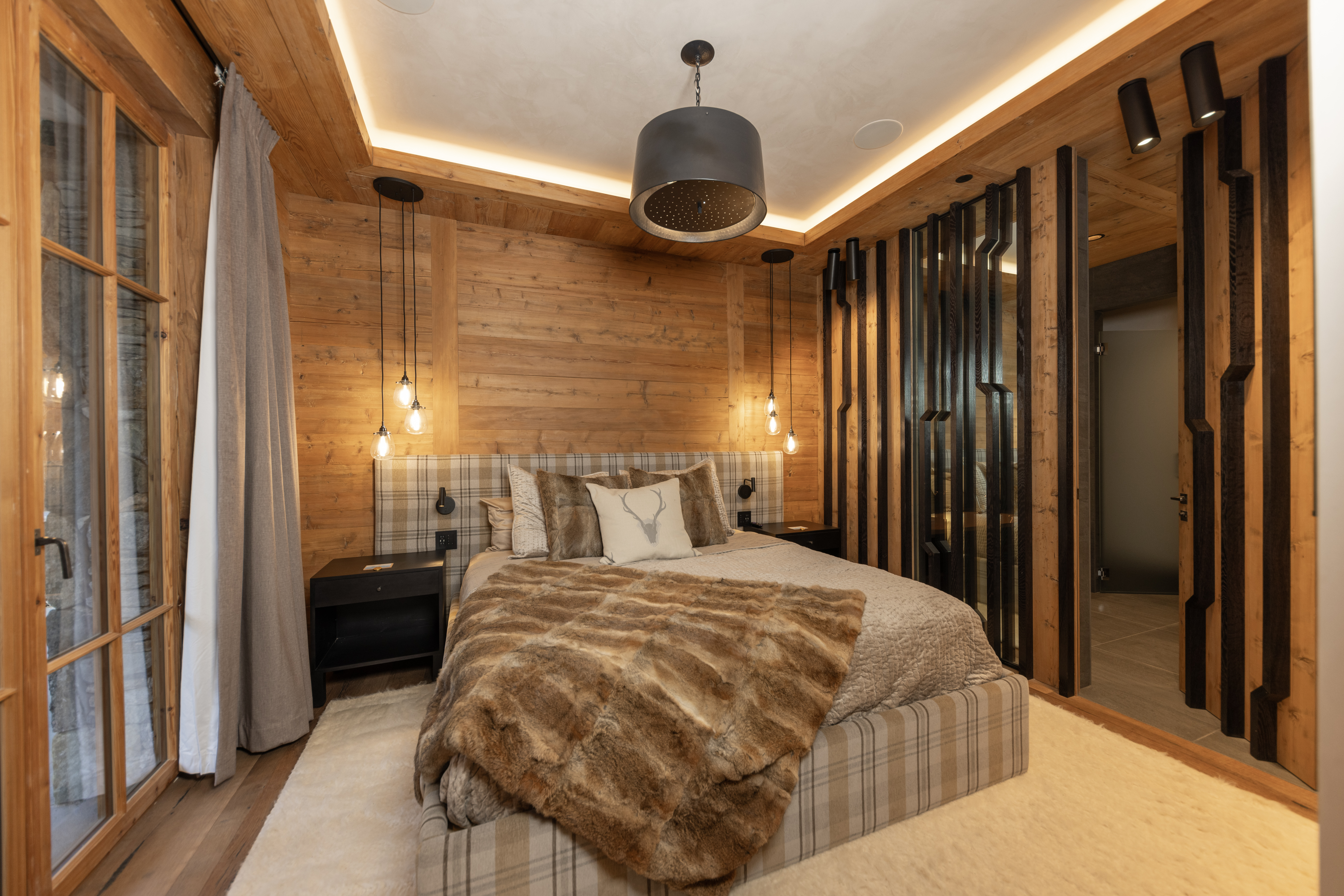 This is a cozy primary bedroom featuring wood paneled walls and ceiling with recessed lighting. The bed has a plaid headboard and is covered with a faux fur throw. The room also includes modern pendant lighting and a decorative wooden partition, creating a warm and inviting atmosphere.