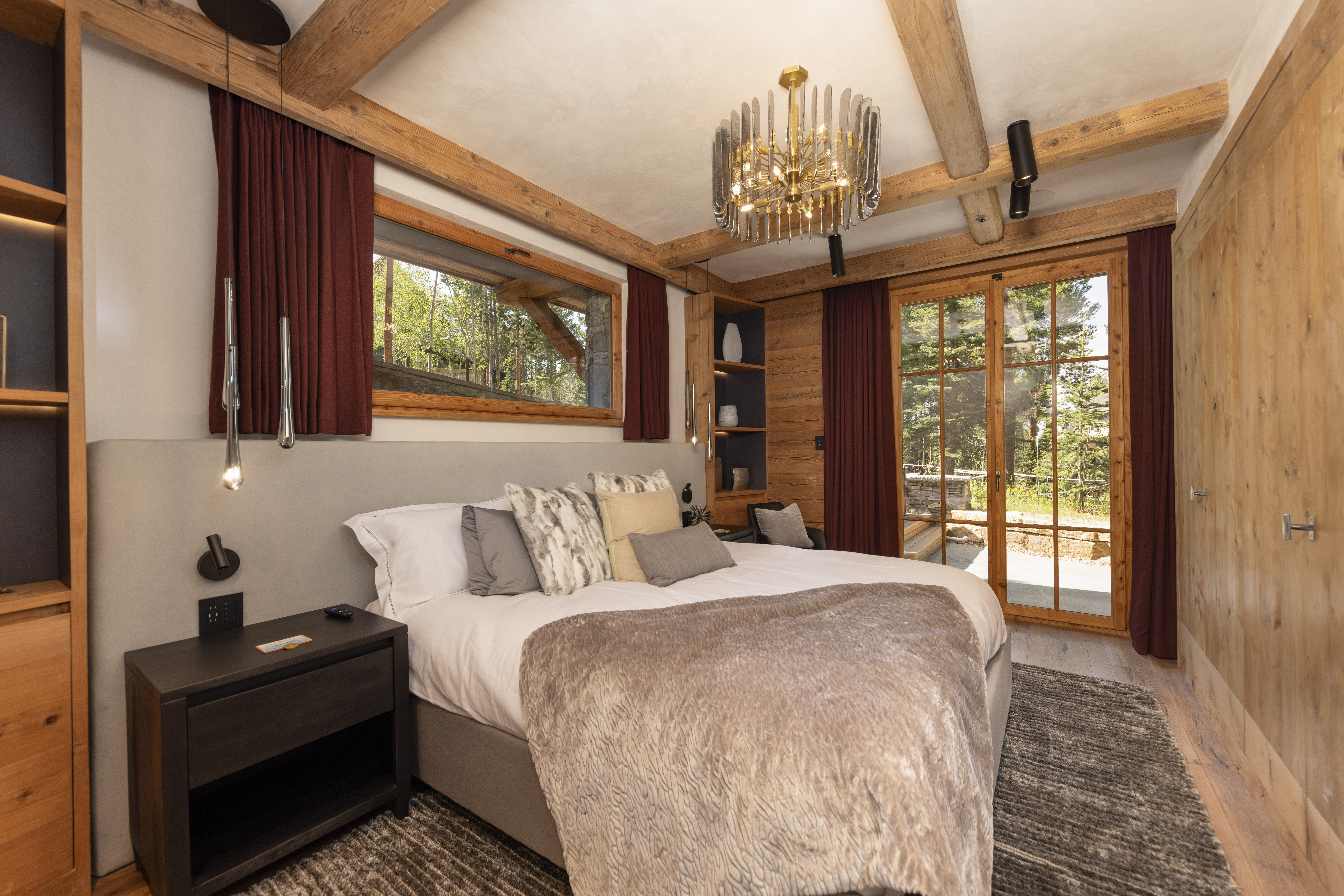 This is a primary bedroom featuring a large bed with multiple pillows and a throw blanket. The room has wooden beams on the ceiling and wood paneling on the walls, creating a rustic yet luxurious feel. A window offers a view of the outdoors, and a modern chandelier hangs above the bed.