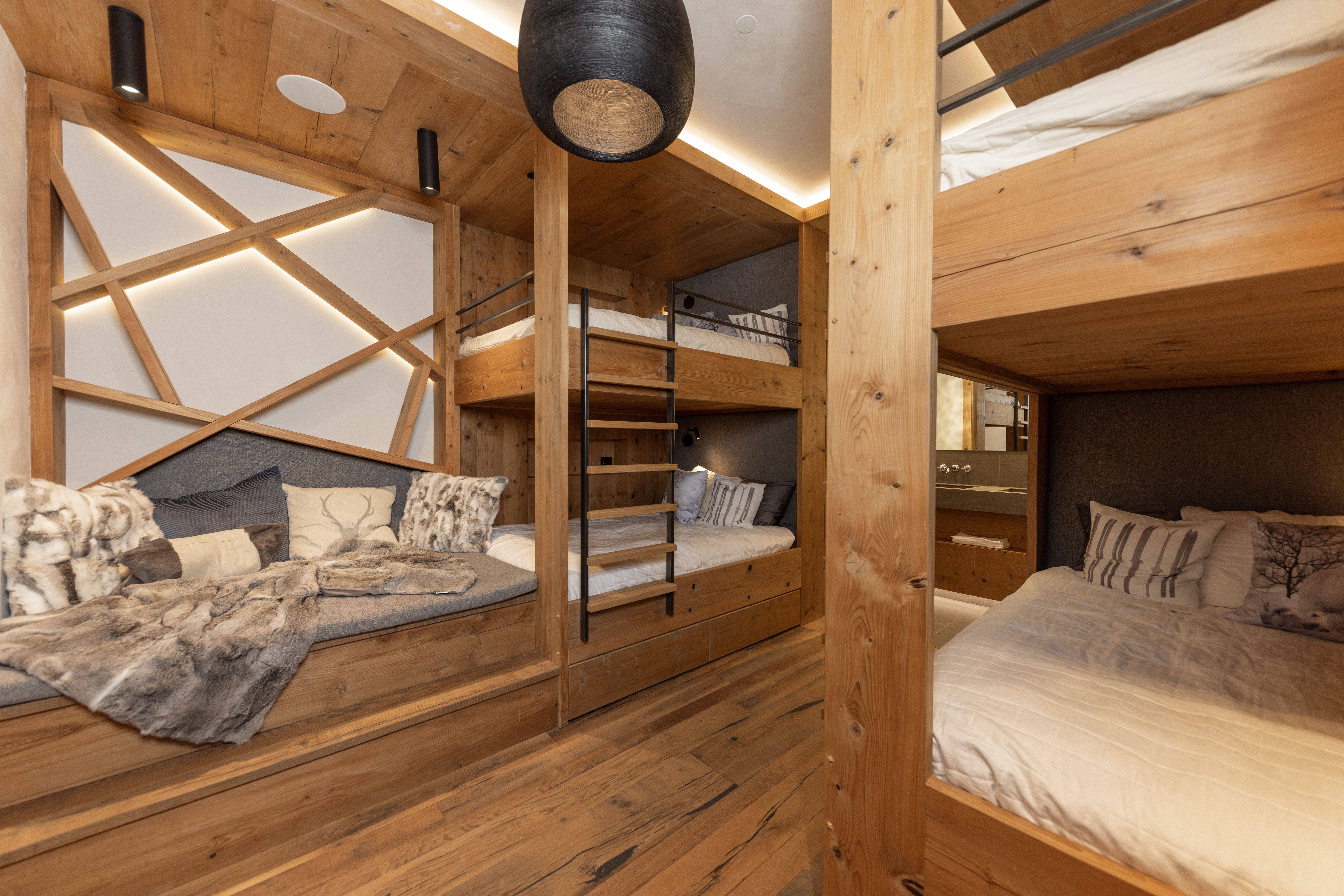 This is an inviting interior shot of a bedroom featuring custom-built wooden bunk beds and a cozy window seat. The room is designed with a rustic, cabin-like aesthetic, highlighted by the natural wood finishes and soft, ambient lighting. The bunk beds offer a practical and stylish sleeping solution, while the window seat provides a comfortable lounging area, making it an ideal space for guests or children.