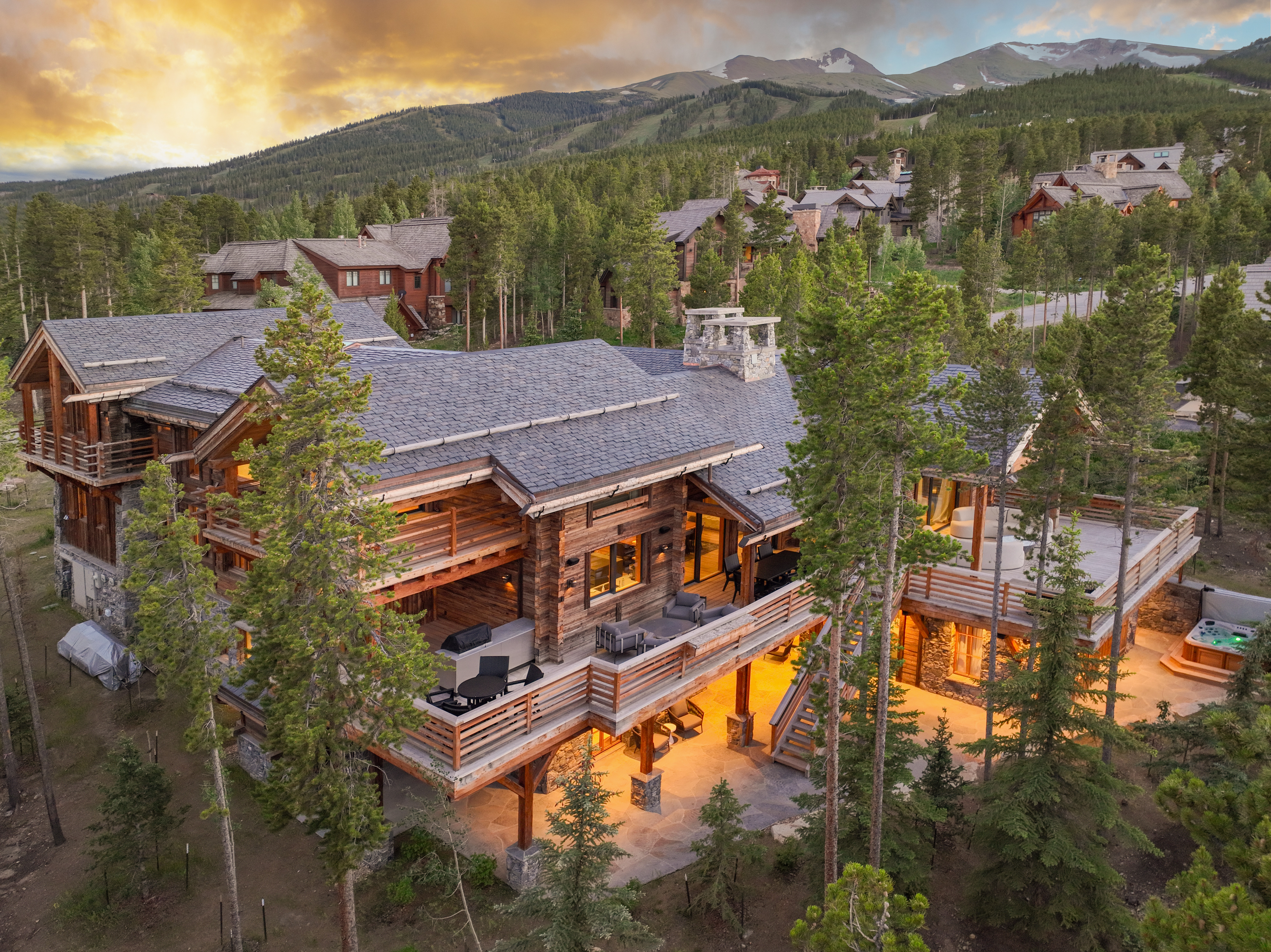 This aerial view showcases a luxurious log cabin home nestled among tall pine trees, with a backdrop of mountains and a vibrant sunset sky. The home features multiple levels of decks and patios, a stone chimney, and a hot tub, creating an inviting outdoor living space. The surrounding landscape emphasizes the property's secluded and natural setting.