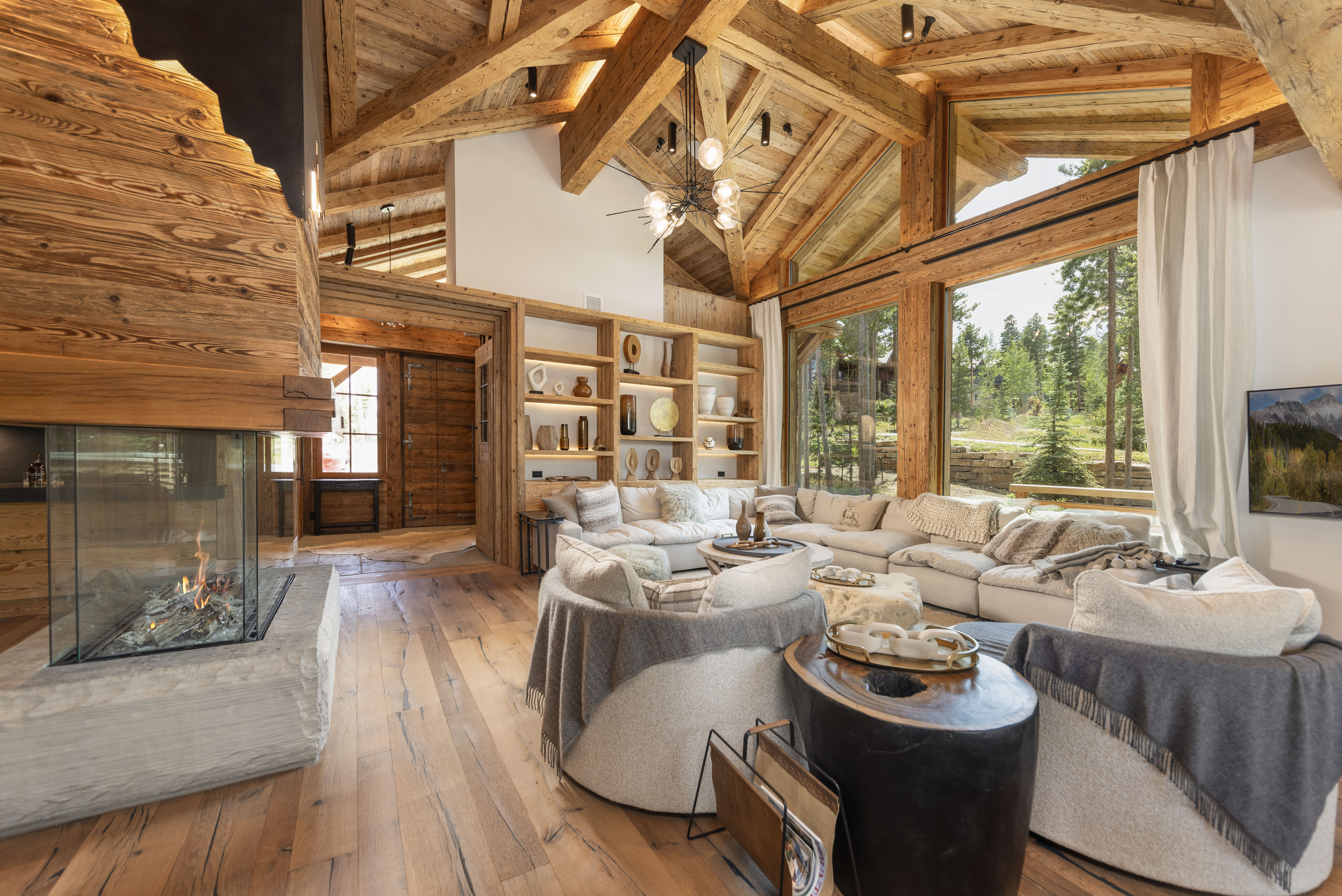 This is an interior shot of a luxurious living room featuring a high, exposed-beam wooden ceiling and a stone fireplace. The room is furnished with a large, comfortable sectional sofa, round ottomans, and built-in shelving displaying decorative items. Large windows offer a view of the surrounding landscape, creating a warm and inviting atmosphere.