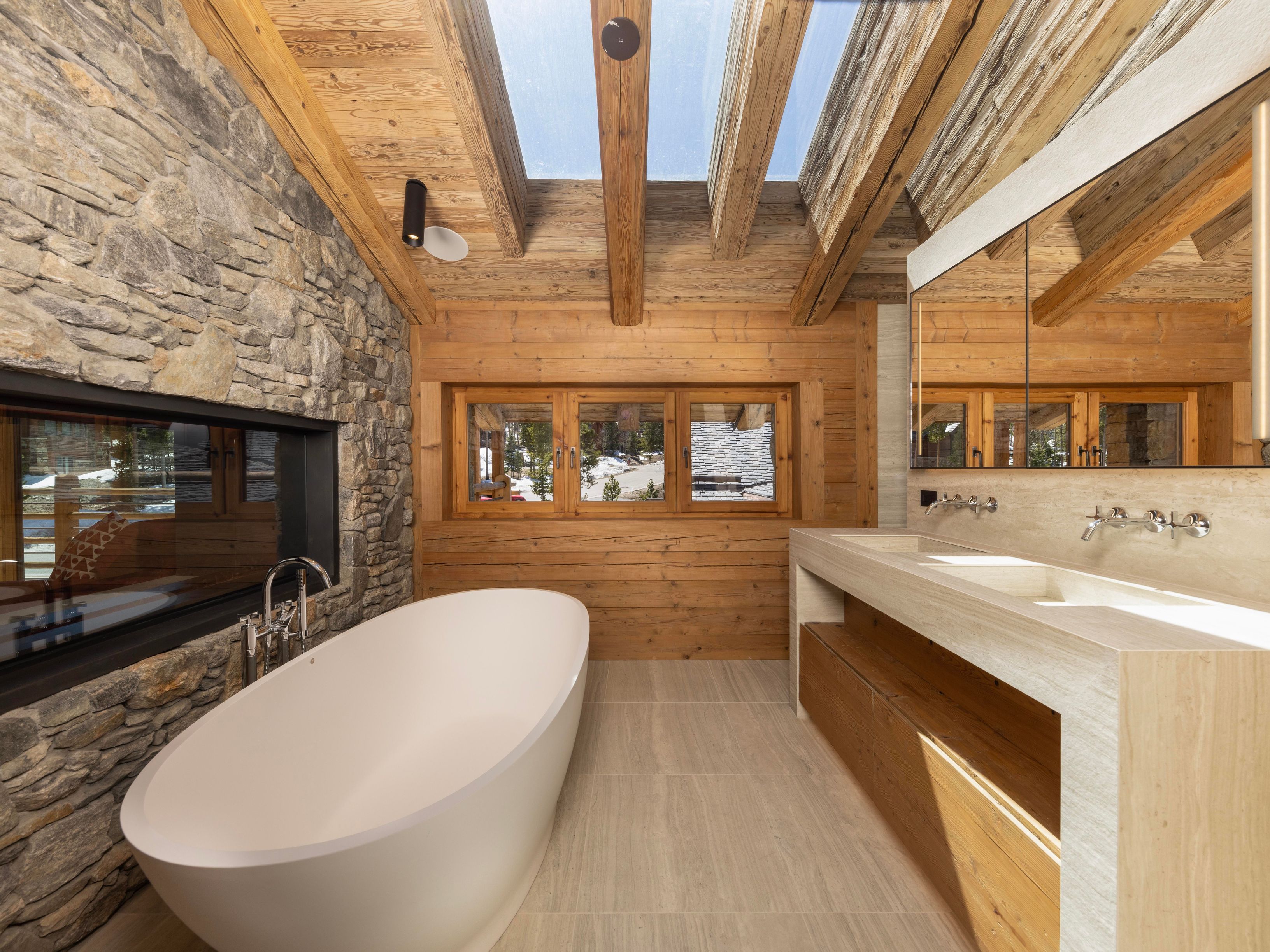 This is a luxurious primary bathroom featuring a freestanding white bathtub positioned next to a stone accent wall with a modern fireplace. The room boasts natural light from a skylight and windows, highlighting the wood-paneled walls and ceiling beams. A double vanity with a light-colored countertop and integrated sinks adds to the spa-like ambiance.
