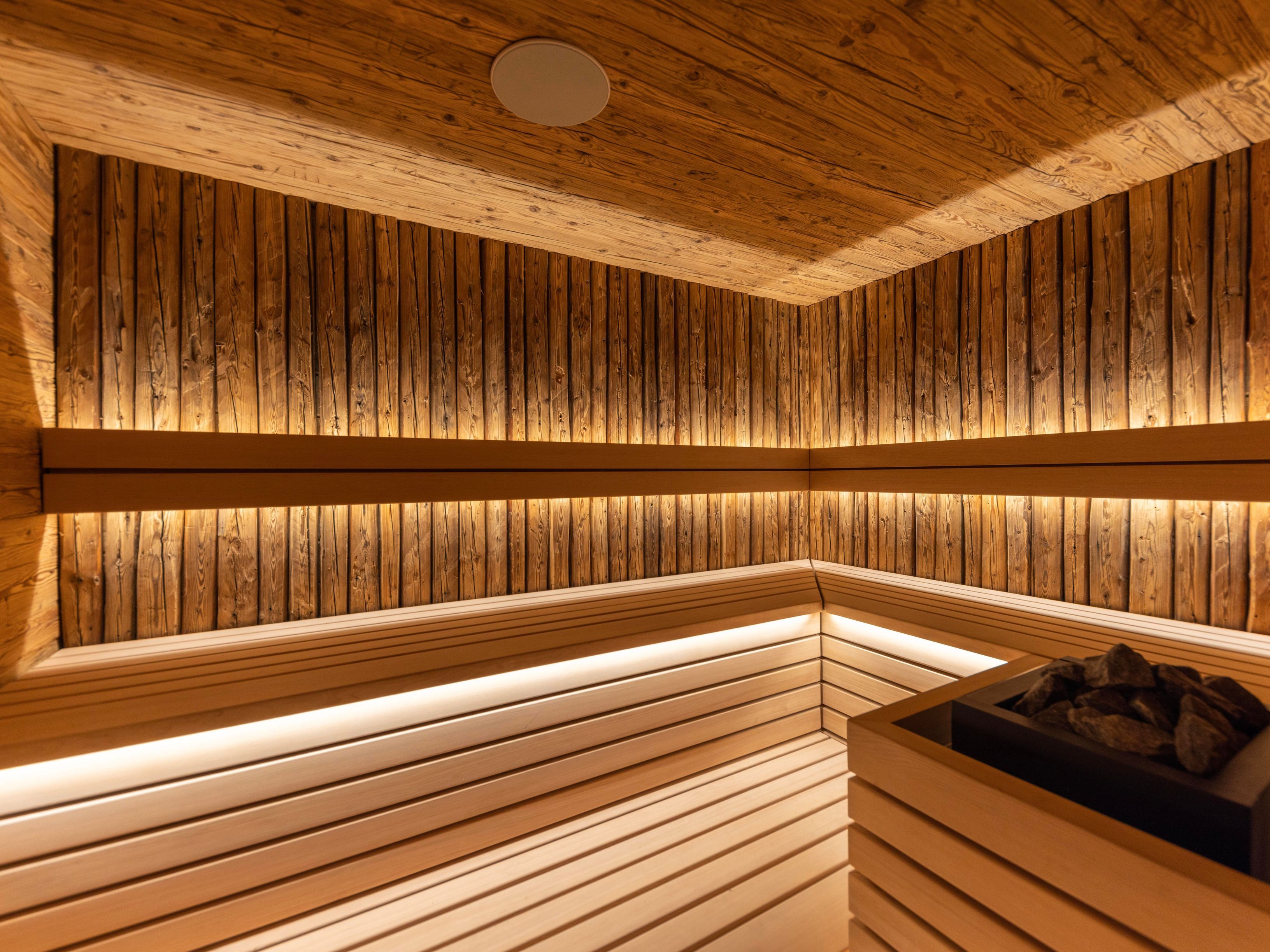 This is an interior shot of a sauna, featuring wooden walls and benches. The wood has a warm, natural tone, and the lighting is soft and inviting, creating a relaxing atmosphere. A heater with rocks is visible in the corner, suggesting a functional and well-maintained space.