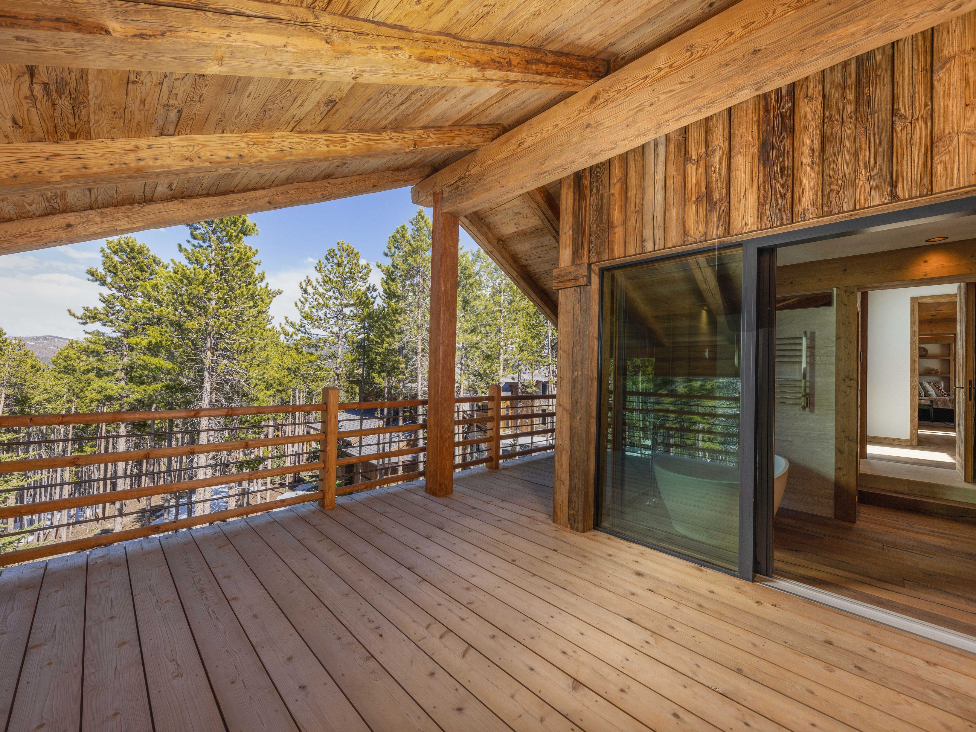 This image showcases a spacious wooden deck or balcony with a rustic, luxurious feel. The deck features a wooden railing and offers views of the surrounding trees and landscape. A large sliding glass door provides access to an interior space, possibly a bathroom, enhancing the indoor-outdoor flow and highlighting the property's connection with nature.
