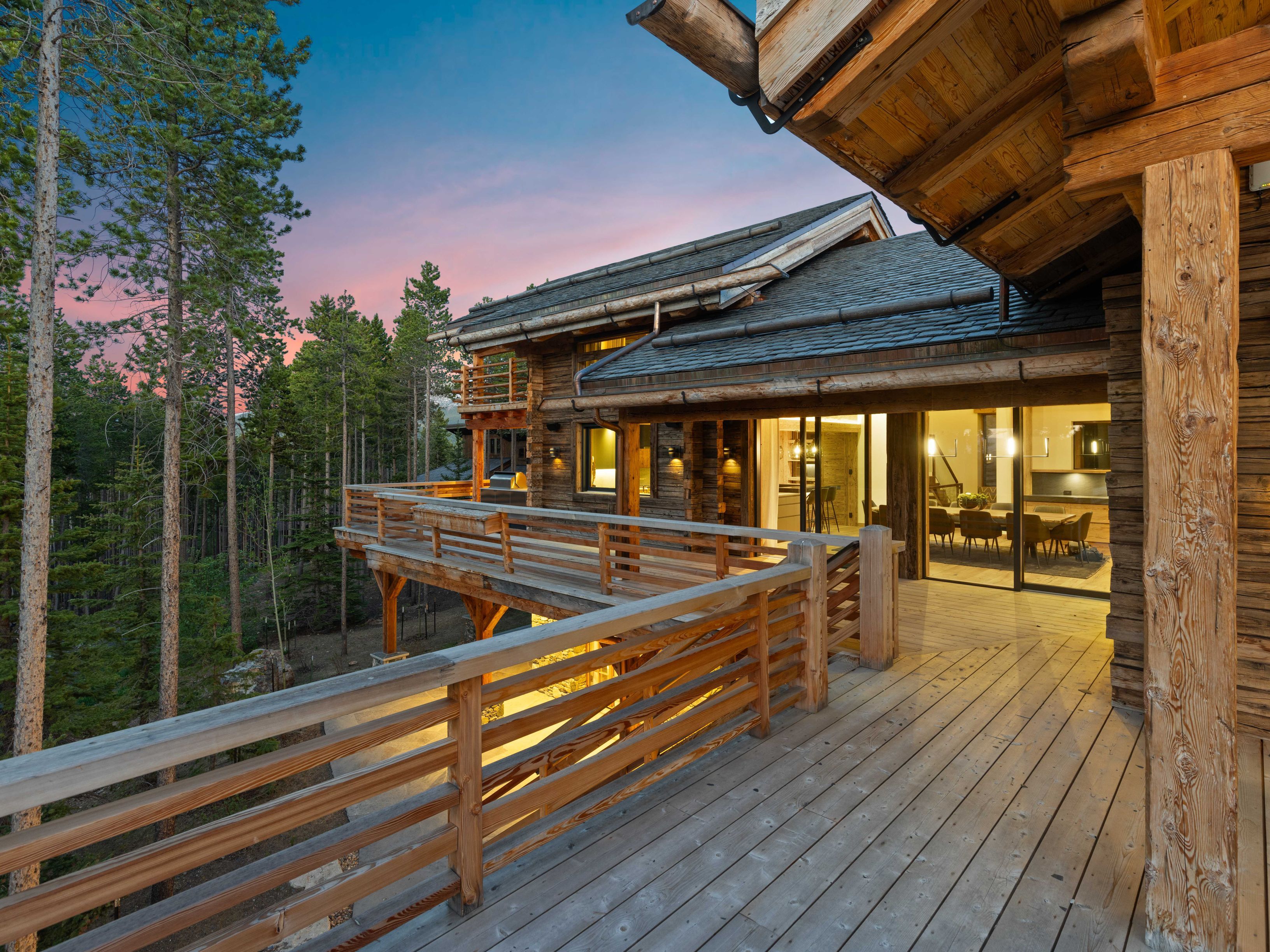 This image showcases a luxurious wooden deck and balcony area of a mountain home at dusk. The deck features wooden railings and connects to the interior through large glass doors, offering a glimpse into the dining area. The surrounding forest and the warm lighting create an inviting and serene atmosphere, highlighting the property's connection to nature.