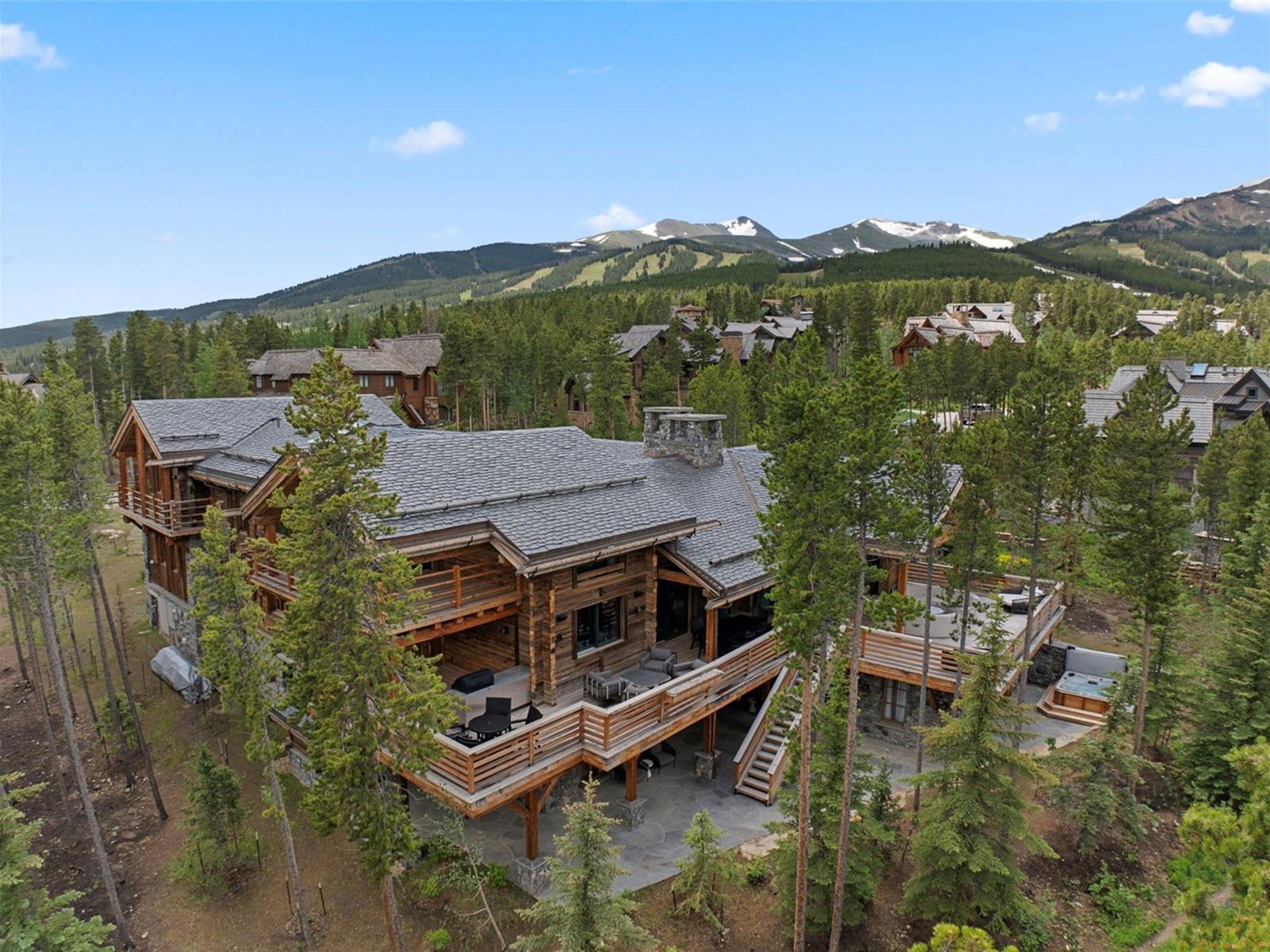 This aerial view showcases a luxurious log cabin home nestled among tall pine trees, with a backdrop of mountains partially covered in snow. The home features multiple decks, a hot tub, and a stone chimney, blending rustic charm with modern amenities. The surrounding landscape includes other high-end homes, suggesting a desirable and exclusive community.