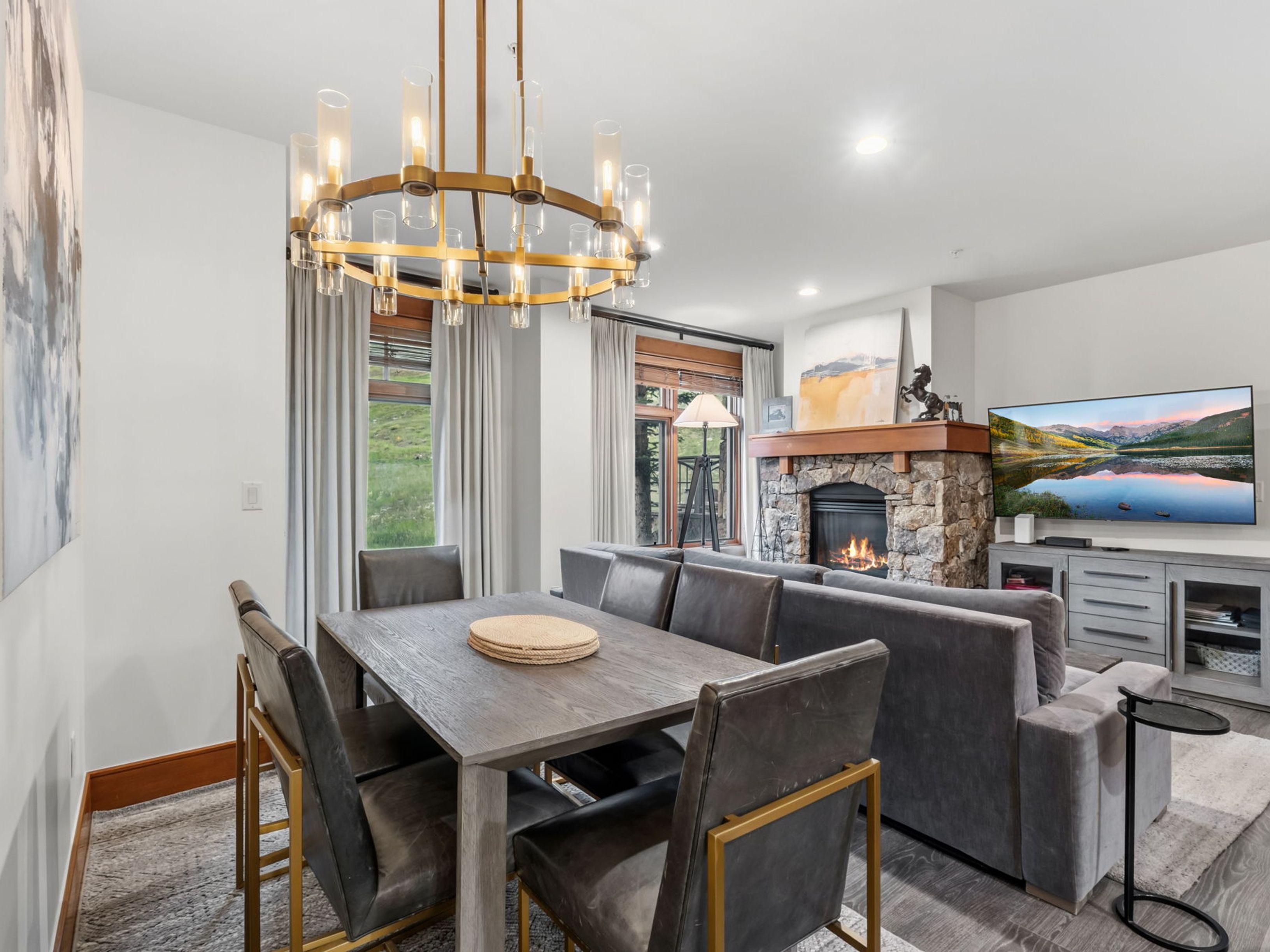 This open-concept living and dining area features a contemporary aesthetic with a dark wood dining table surrounded by leather chairs with gold-toned metal frames. A large, circular chandelier serves as a focal point above the dining space, while the adjacent living area includes a plush grey sofa and a stone-clad fireplace with a wooden mantel. The room is brightened by large windows with neutral curtains, creating a warm and inviting atmosphere suitable for both entertaining and relaxation.