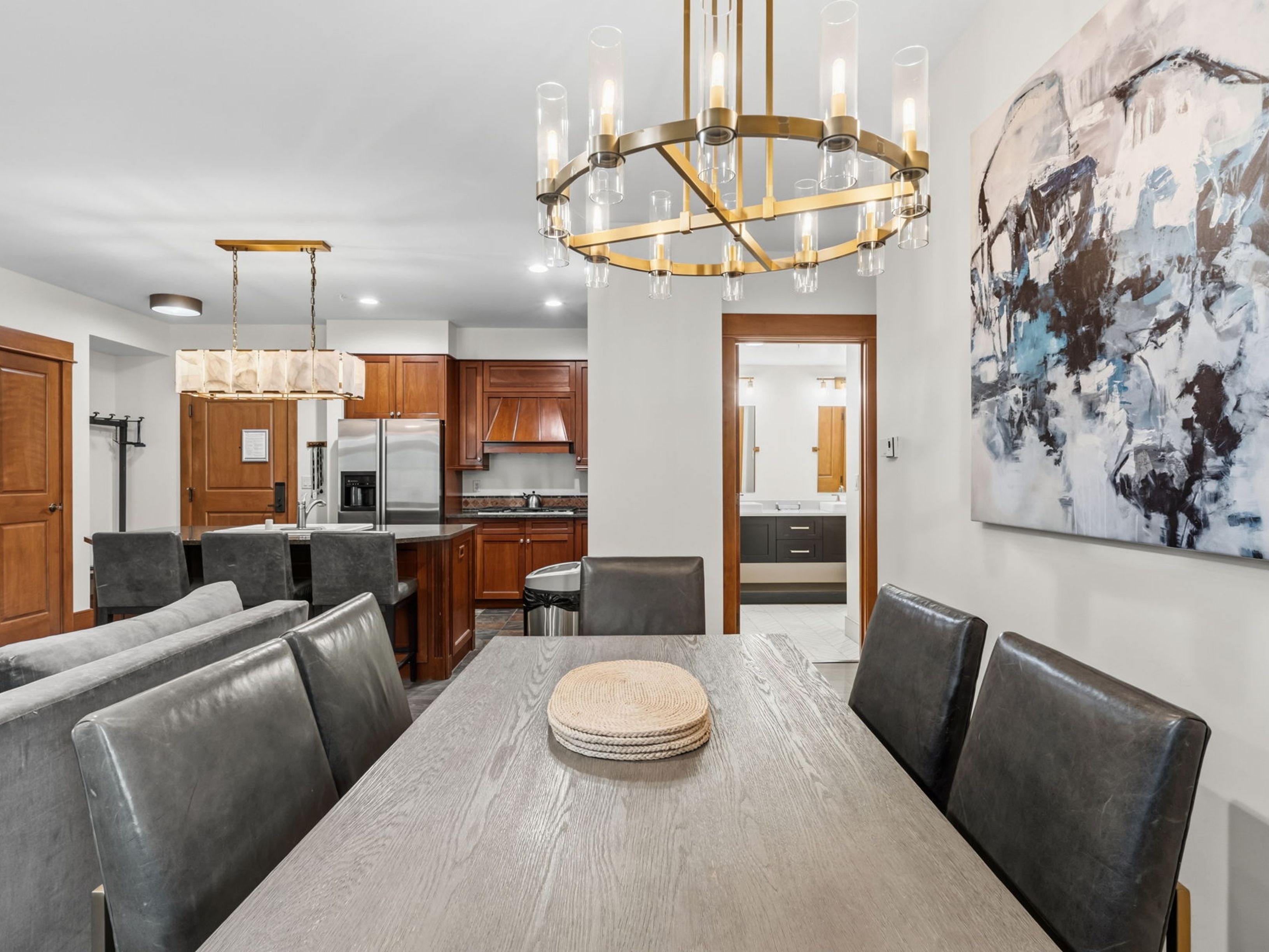 This open-concept dining area features a large, light-toned wooden table surrounded by dark leather-upholstered chairs, positioned beneath a prominent, multi-tiered brass chandelier. The space flows seamlessly into a kitchen with warm-toned cabinetry and a breakfast bar, while a large abstract painting adds a vibrant focal point to the dining wall. A doorway in the background provides a glimpse into a secondary bathroom, highlighting the home's cohesive and functional layout.