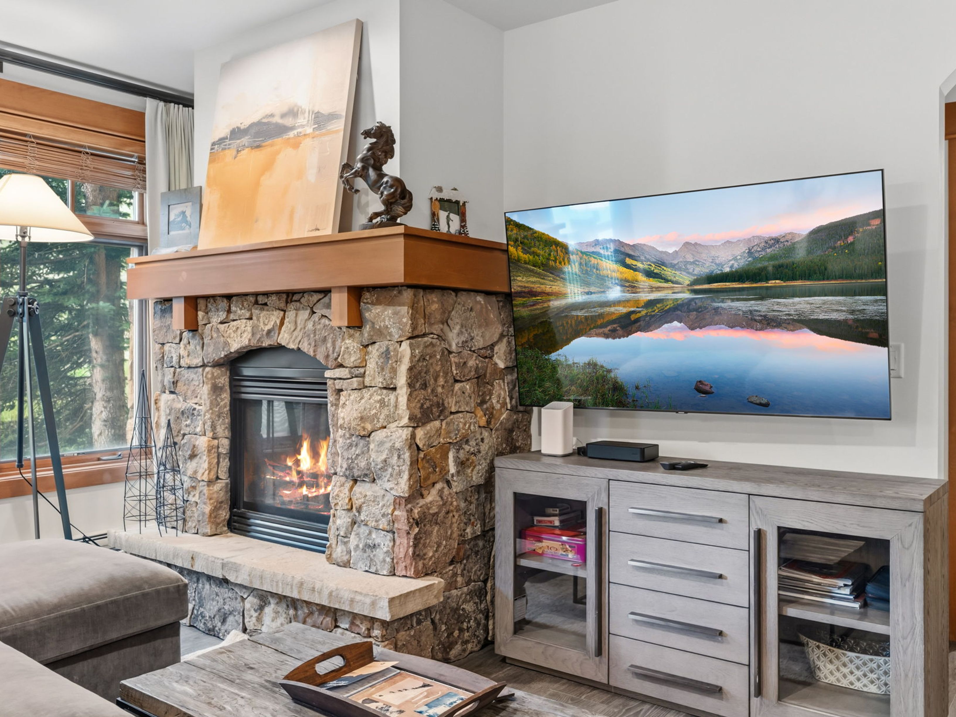 This cozy living room features a prominent stone-clad fireplace with a wooden mantel, serving as a rustic focal point. To the right, a large flat-screen television is mounted above a light-toned wooden media console, while a window to the left provides natural light and a view of the outdoors. The space is furnished with a comfortable sofa and a wooden coffee table, creating a warm and inviting atmosphere.