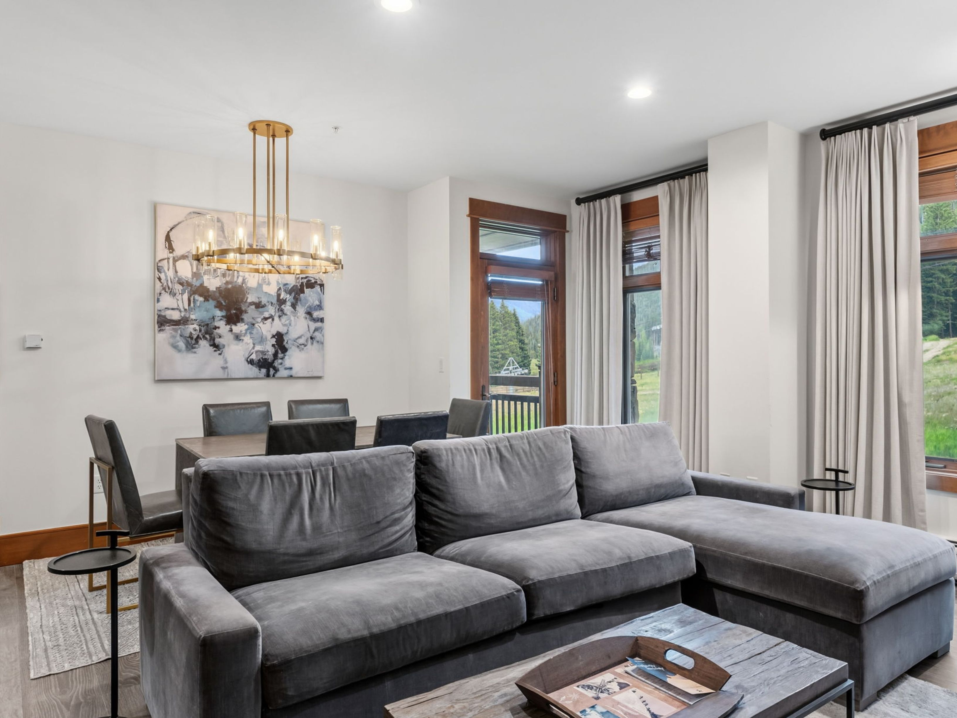 This open-concept living and dining area features a large, plush grey sectional sofa in the foreground and a dining table with six chairs in the background. A modern gold-toned chandelier hangs above the dining space, complemented by abstract wall art and tall windows that provide a view of the surrounding forest. The room is finished with light-toned wood flooring and neutral walls, creating a bright and inviting atmosphere.