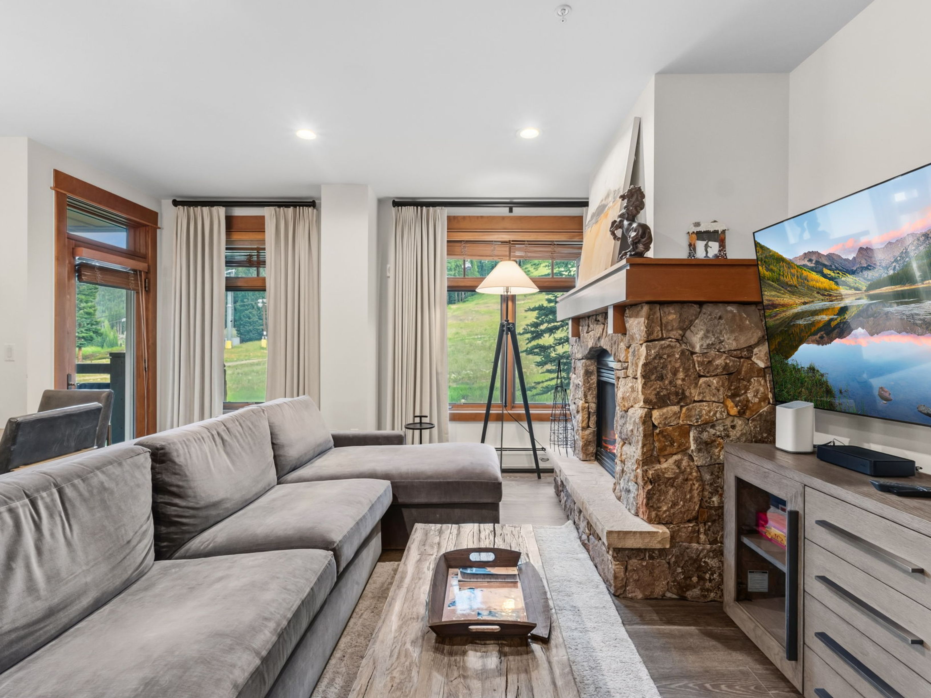 This cozy living room features a large, plush grey sectional sofa arranged around a rustic wooden coffee table. A prominent stone-clad chimney breast with a built-in fireplace serves as the focal point, topped by a wooden mantel and decorative art. Large windows with neutral curtains frame a scenic view of a grassy hillside, while a tripod floor lamp adds a touch of modern style to the space.