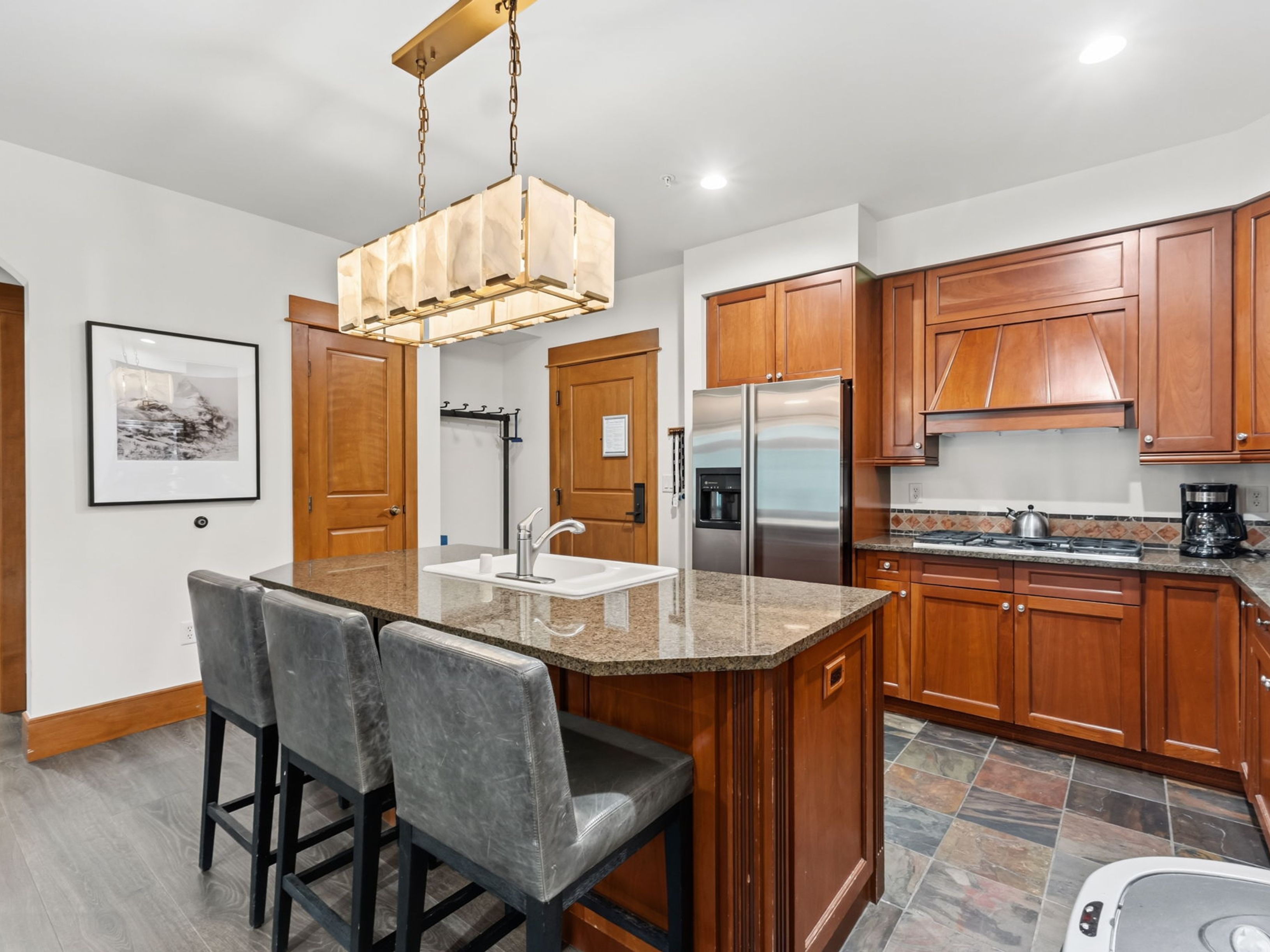 This kitchen features warm-toned wood cabinetry, a central island with a stone countertop and integrated sink, and a multi-light pendant fixture overhead. The space is equipped with stainless steel appliances, including a refrigerator and a gas range, complemented by a multi-colored slate tile floor. Three gray upholstered bar stools provide seating at the island, creating a functional and inviting atmosphere.