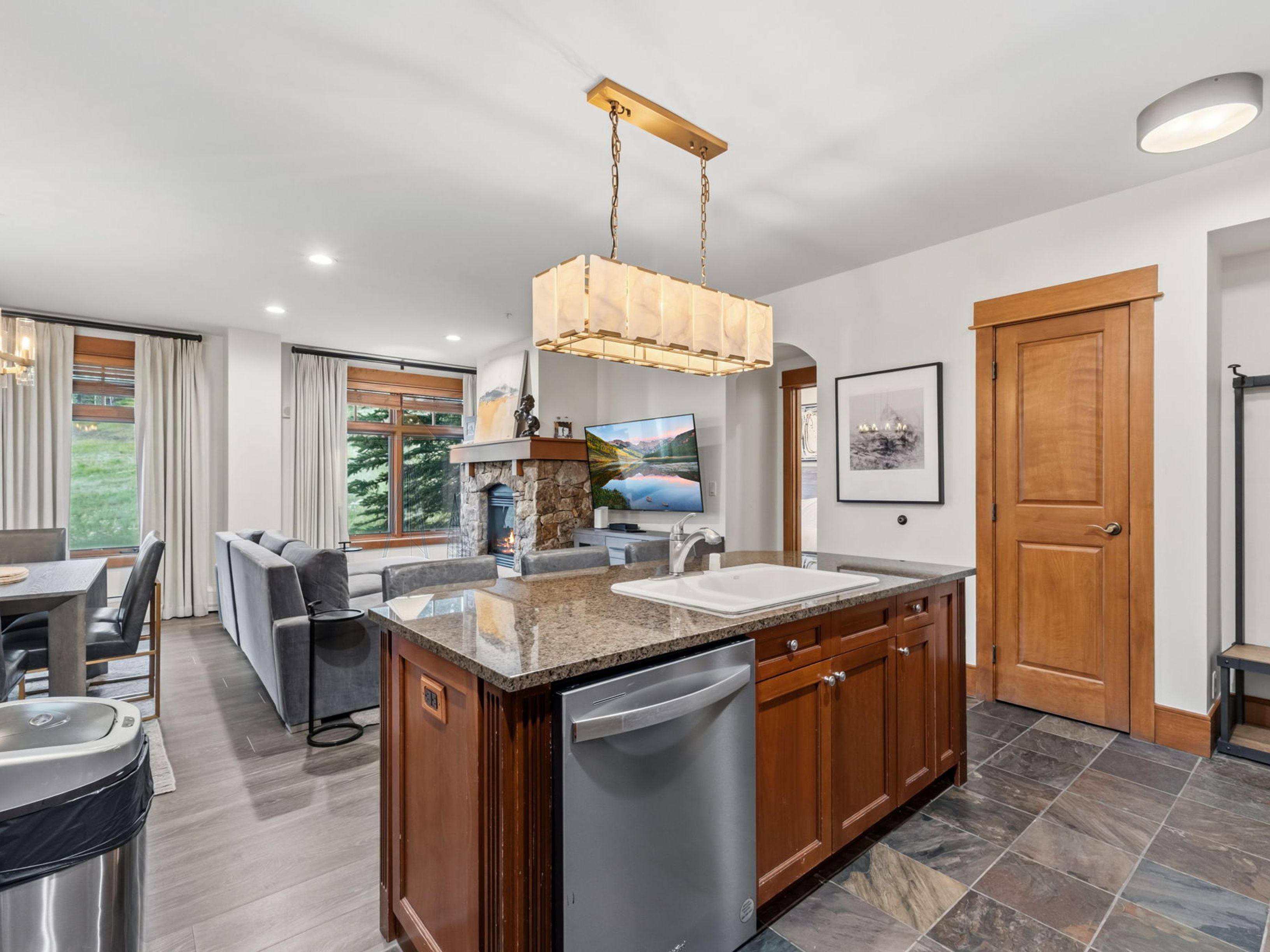 This open-concept kitchen features a central island with a stone countertop, a stainless steel dishwasher, and a white sink, seamlessly flowing into the adjacent living area. The space is defined by warm wood cabinetry, a distinctive rectangular pendant light fixture, and slate-style floor tiles. A stone-clad chimney breast serves as a focal point in the living room, which is bathed in natural light from large windows overlooking the outdoors.