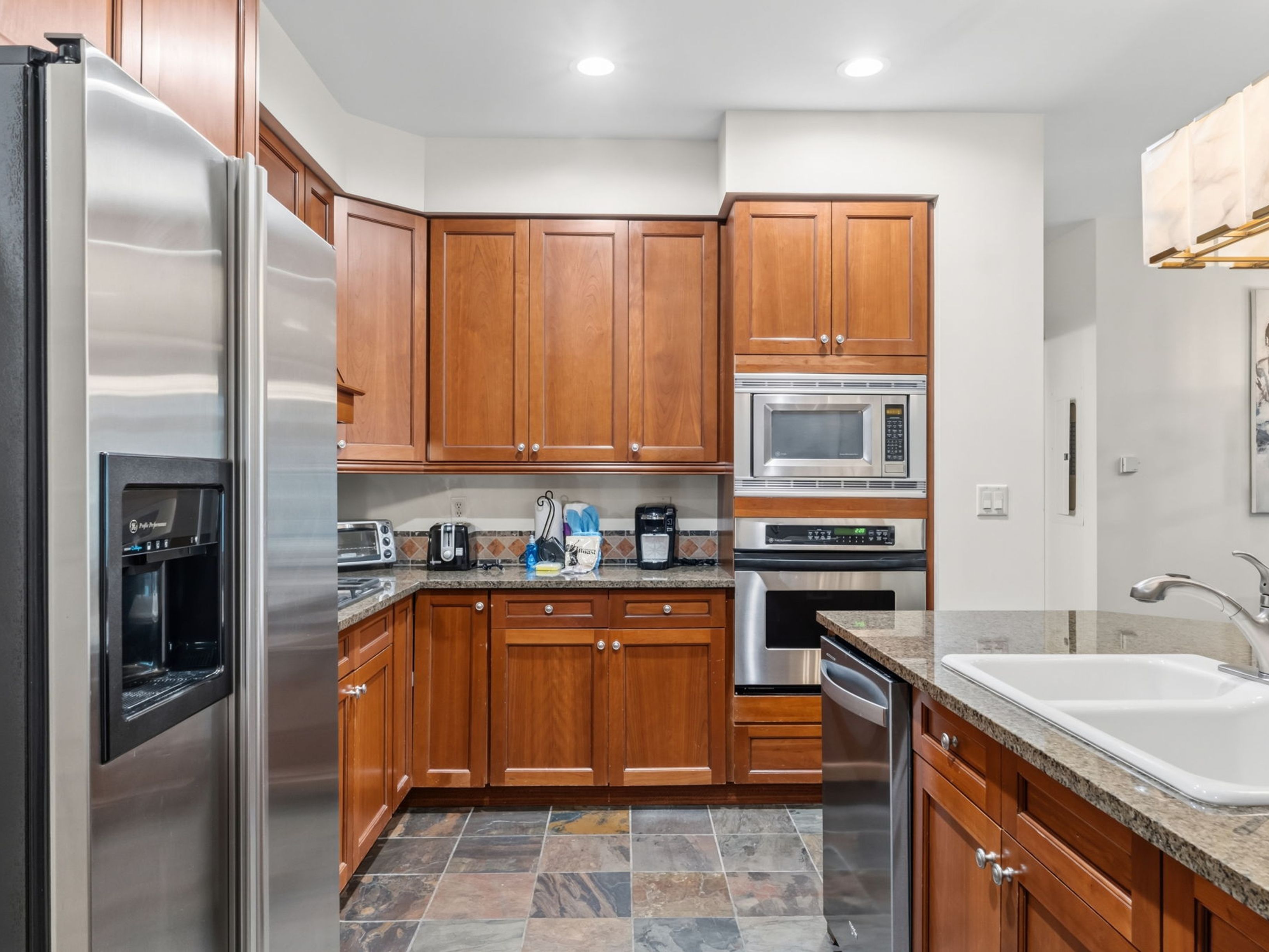 This kitchen features warm-toned wood cabinetry paired with speckled stone countertops and a multi-colored slate tile floor. The space is equipped with stainless steel appliances, including a side-by-side refrigerator, a built-in microwave, and an oven. A peninsula with a white double-basin sink extends into the foreground, creating a functional layout for cooking and meal preparation.