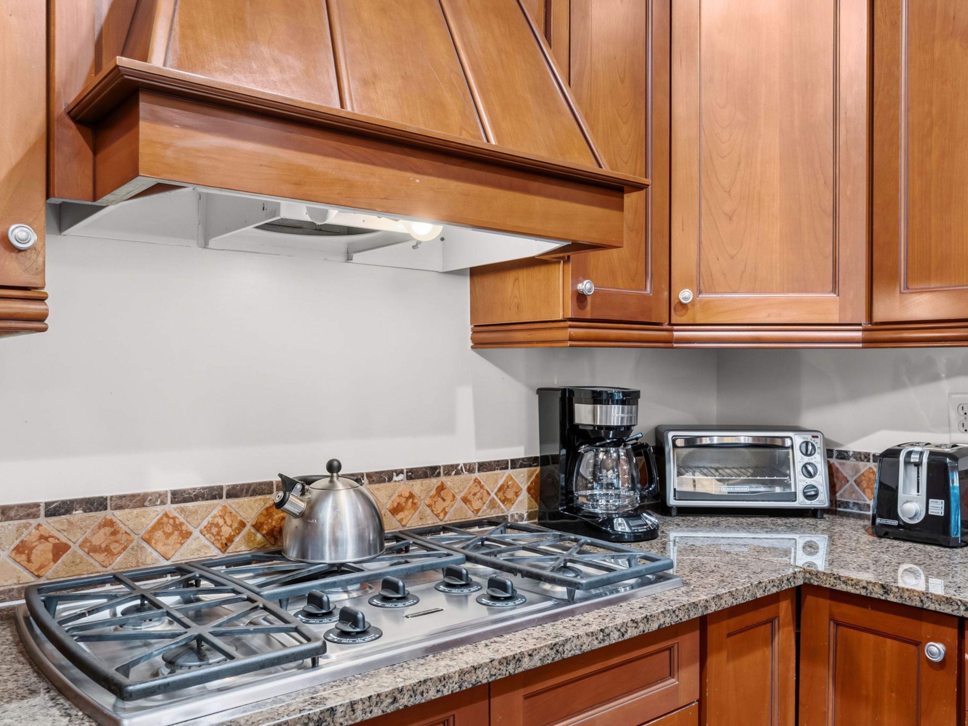 This kitchen detail shot highlights a stainless steel gas cooktop set into a speckled stone countertop, complemented by a metallic tea kettle. Above the stove, a custom wood-paneled range hood sits against a neutral wall, accented by a decorative backsplash of diamond-patterned tiles. To the right, a coffee maker and toaster are neatly arranged on the counter, framed by warm-toned wooden cabinetry.