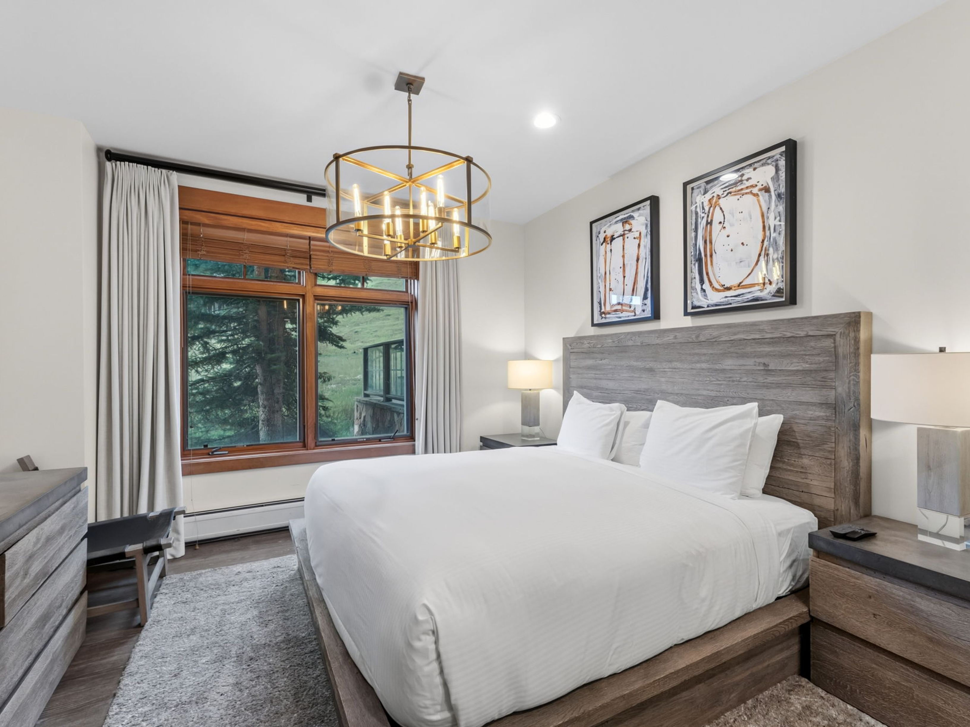 This primary bedroom features a rustic-modern aesthetic with a large wooden platform bed as the centerpiece, complemented by matching wooden nightstands and a dresser. A prominent circular chandelier hangs from the ceiling, while two abstract framed artworks are mounted above the headboard. Large windows with neutral curtains provide a view of the surrounding trees and landscape, creating a serene and inviting atmosphere.