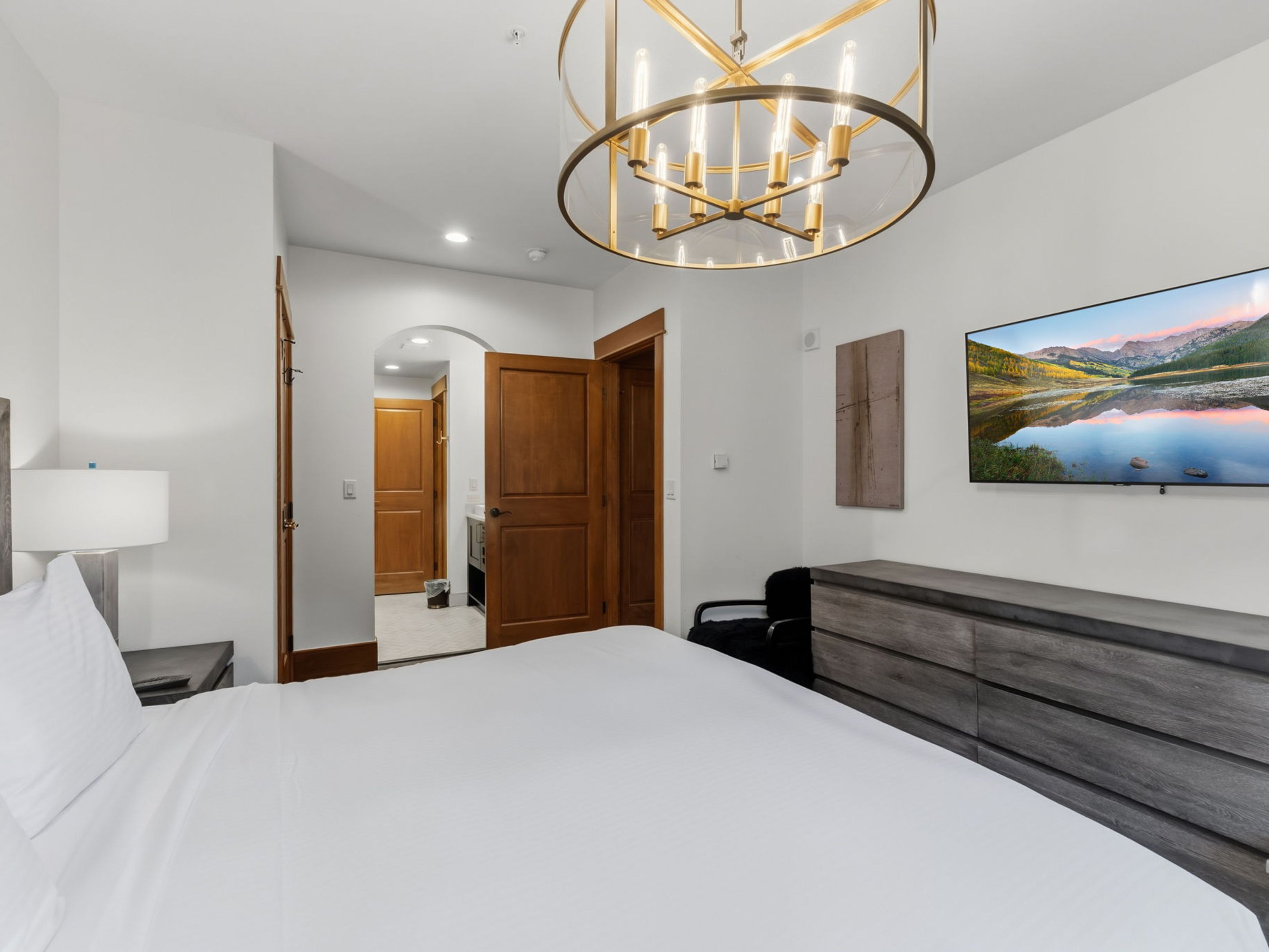 This primary bedroom features a crisp, white-themed aesthetic with a large bed as the central focus. A prominent gold-toned circular chandelier hangs from the ceiling, while a wall-mounted television sits above a dark wood dresser. The room includes an arched doorway leading to an adjoining bathroom, showcasing a clean and well-lit layout.