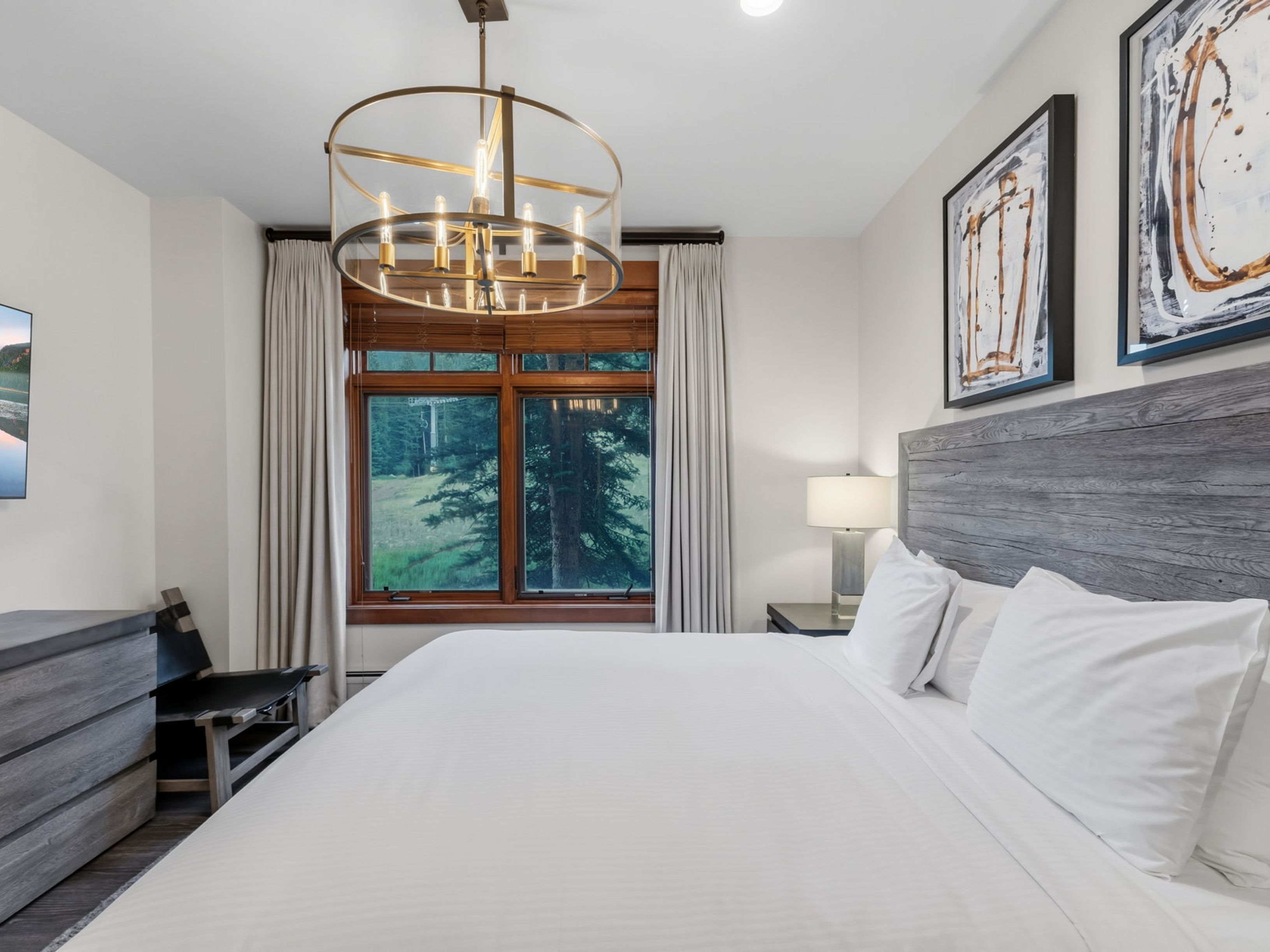 This primary bedroom features a large bed with a rustic, weathered-wood headboard and crisp white bedding, centered in the room. A prominent brass chandelier hangs from the ceiling, while a window with wooden blinds and light curtains provides a view of the surrounding trees. To the left, a matching weathered-wood dresser sits beside a modern chair, and two framed abstract artworks hang above the bed, creating a cohesive, mountain-inspired aesthetic.