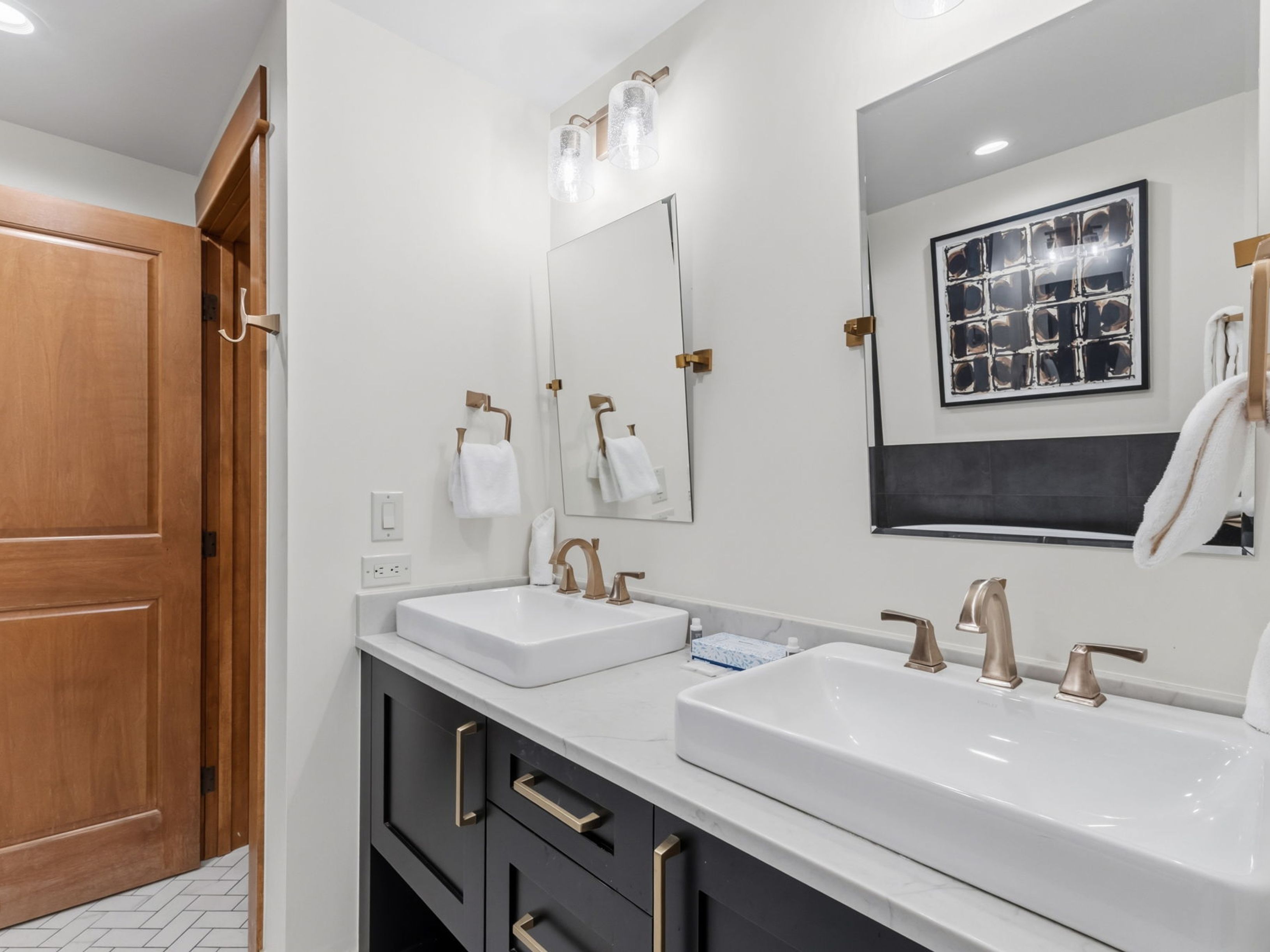 This modern primary bathroom features a double vanity with a dark cabinet base, light stone countertops, and rectangular vessel sinks. The space is accented with warm-toned metal fixtures, including faucets, towel rings, and cabinet hardware, complemented by a pair of rectangular mirrors and glass pendant lighting. A herringbone-patterned floor adds texture to the room, which is finished with crisp white walls and a framed piece of abstract art.