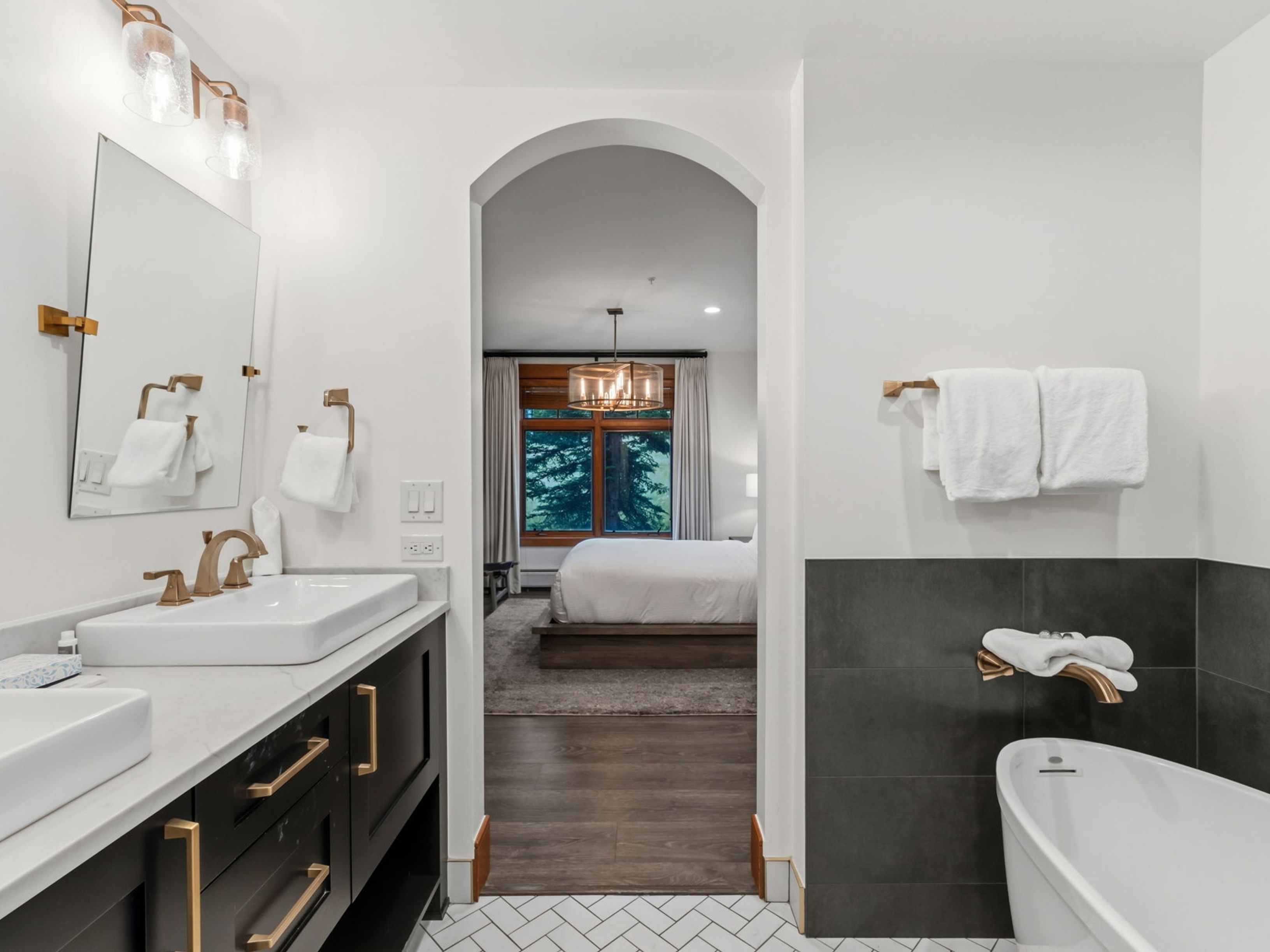 This primary bathroom features a dual-sink vanity with dark cabinetry, gold-toned hardware, and white countertops, complemented by a freestanding soaking tub set against a dark tiled wall. An arched doorway provides a seamless transition into a bright bedroom, creating an open and airy flow between the spaces. The room is finished with a herringbone-patterned floor, adding a sophisticated touch to the modern design.