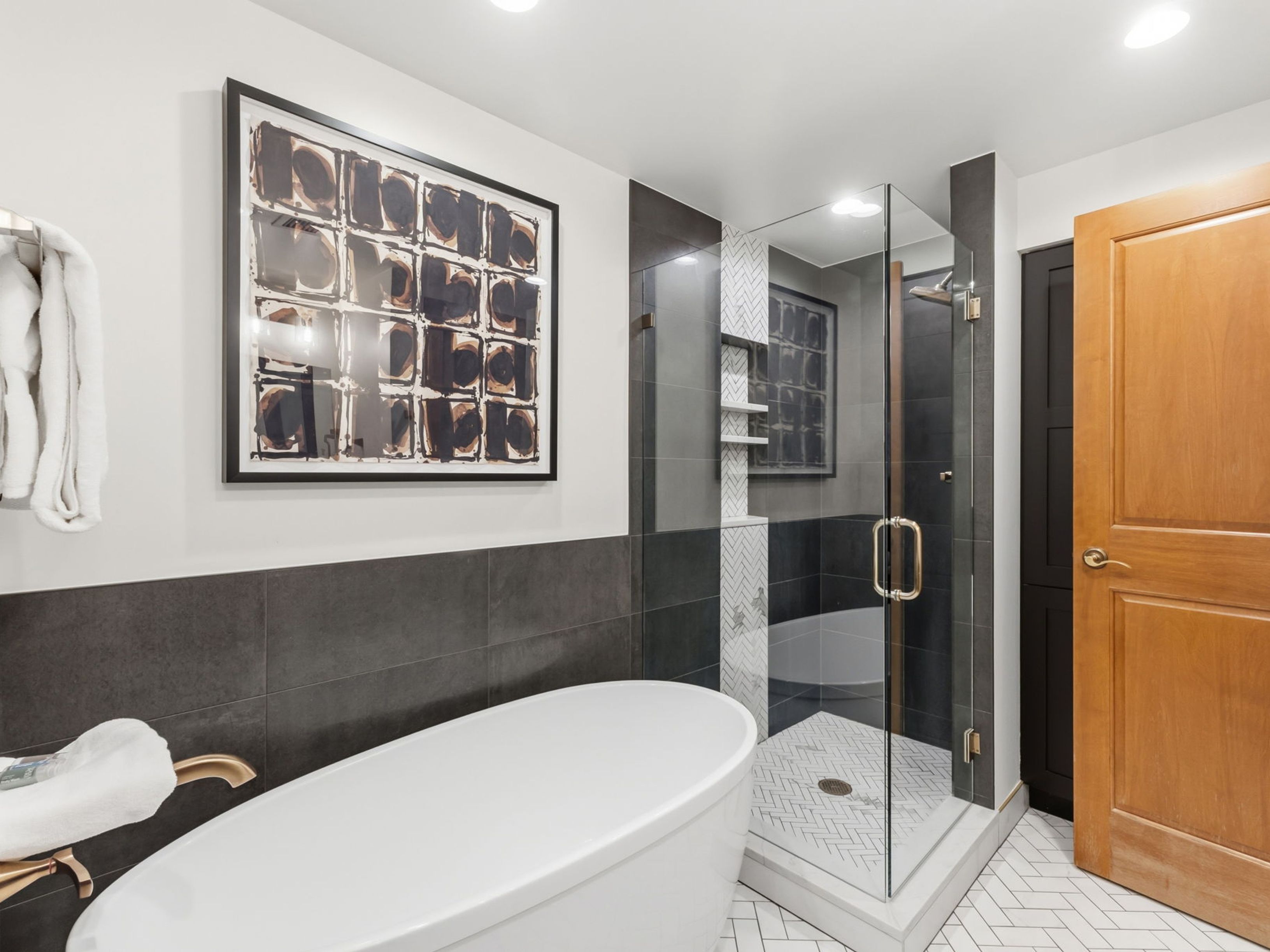 This modern bathroom features a sleek, freestanding white soaking tub paired with a glass-enclosed walk-in shower. The shower is accented with a vertical strip of herringbone-patterned tile and built-in shelving, while the floor showcases a matching herringbone tile design. A large, framed abstract art piece hangs on the wall above the tub, contributing to the sophisticated and contemporary aesthetic of the space.
