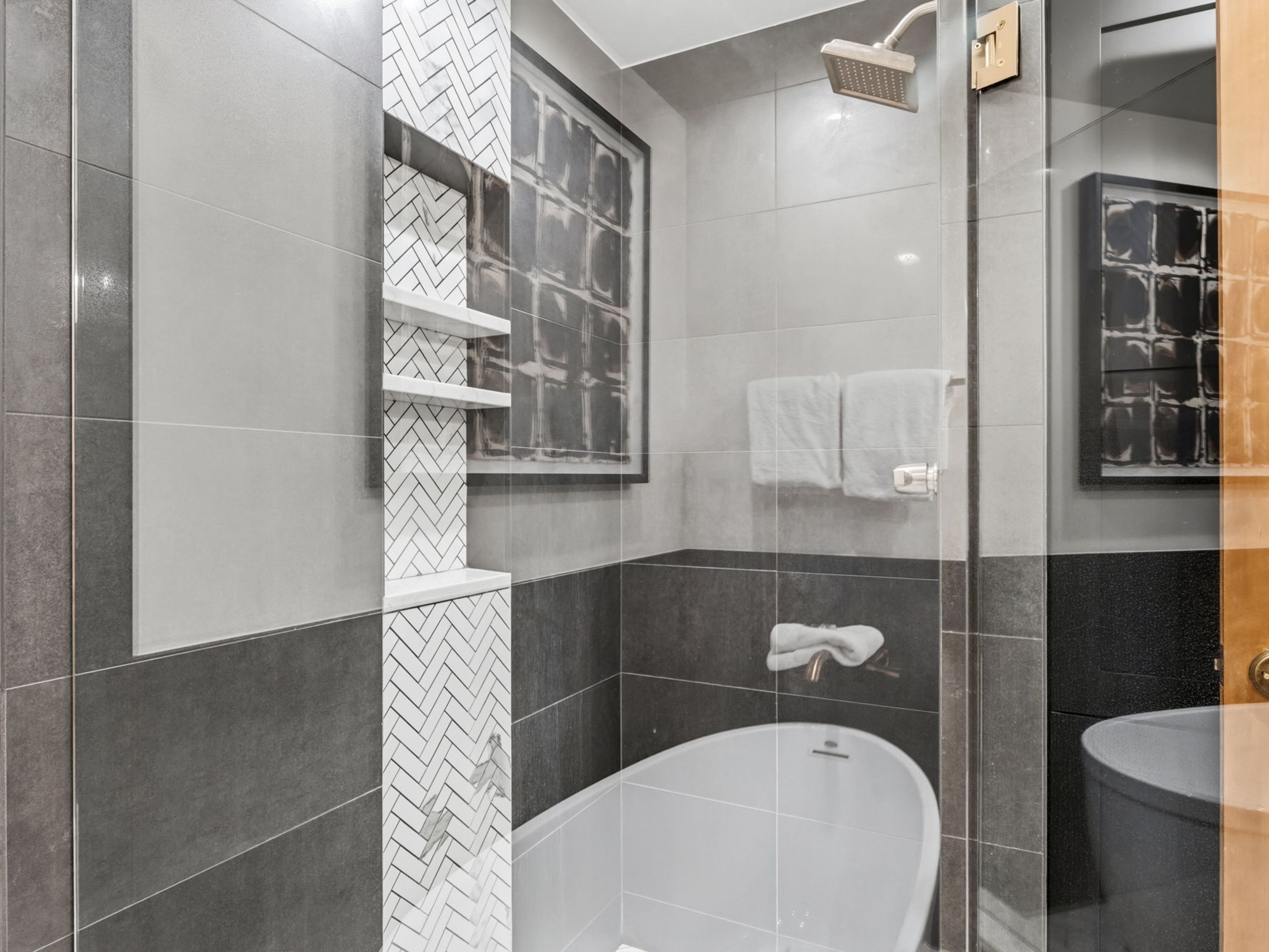 This modern bathroom features a sleek, freestanding soaking tub positioned beneath a shower head, enclosed by a glass partition. A striking vertical accent wall with white herringbone tile incorporates built-in shelving, contrasting with the surrounding large-format gray and dark charcoal tiles. The space is accented by a framed piece of abstract art on the wall, creating a sophisticated and contemporary aesthetic.