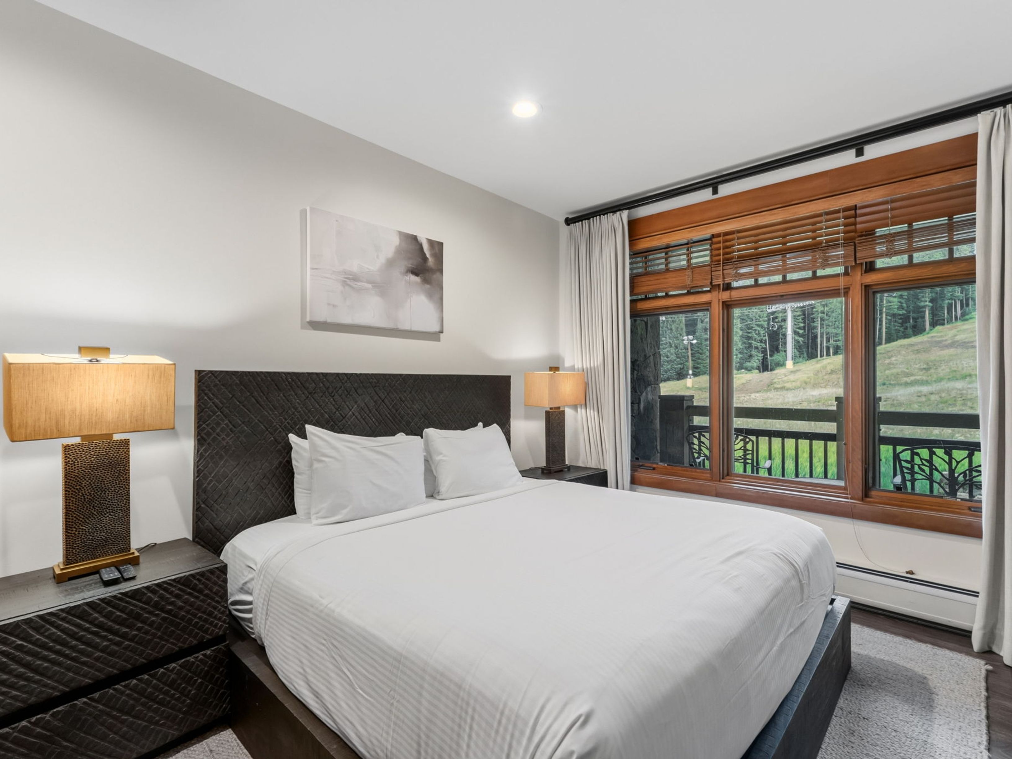 This bedroom features a dark, textured headboard and matching nightstands, creating a sophisticated and cozy atmosphere. A large window with wooden blinds and light curtains offers a scenic view of the ski slopes outside. The room is accented by two warm-toned table lamps and a piece of abstract wall art, providing a comfortable and inviting retreat.