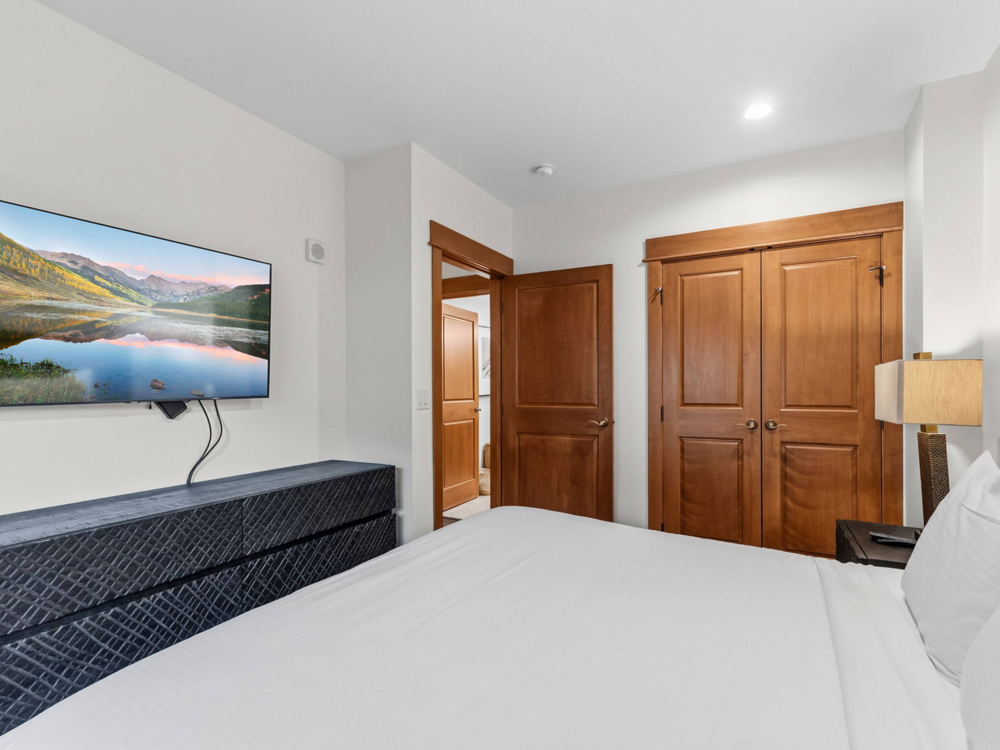 This bedroom features a clean, neutral aesthetic with a white-made bed and a dark, textured dresser positioned beneath a wall-mounted television. A set of wooden double doors leads to a closet, while an open doorway provides a glimpse into an adjacent room. The space is illuminated by a bedside lamp, creating a cozy and functional atmosphere.