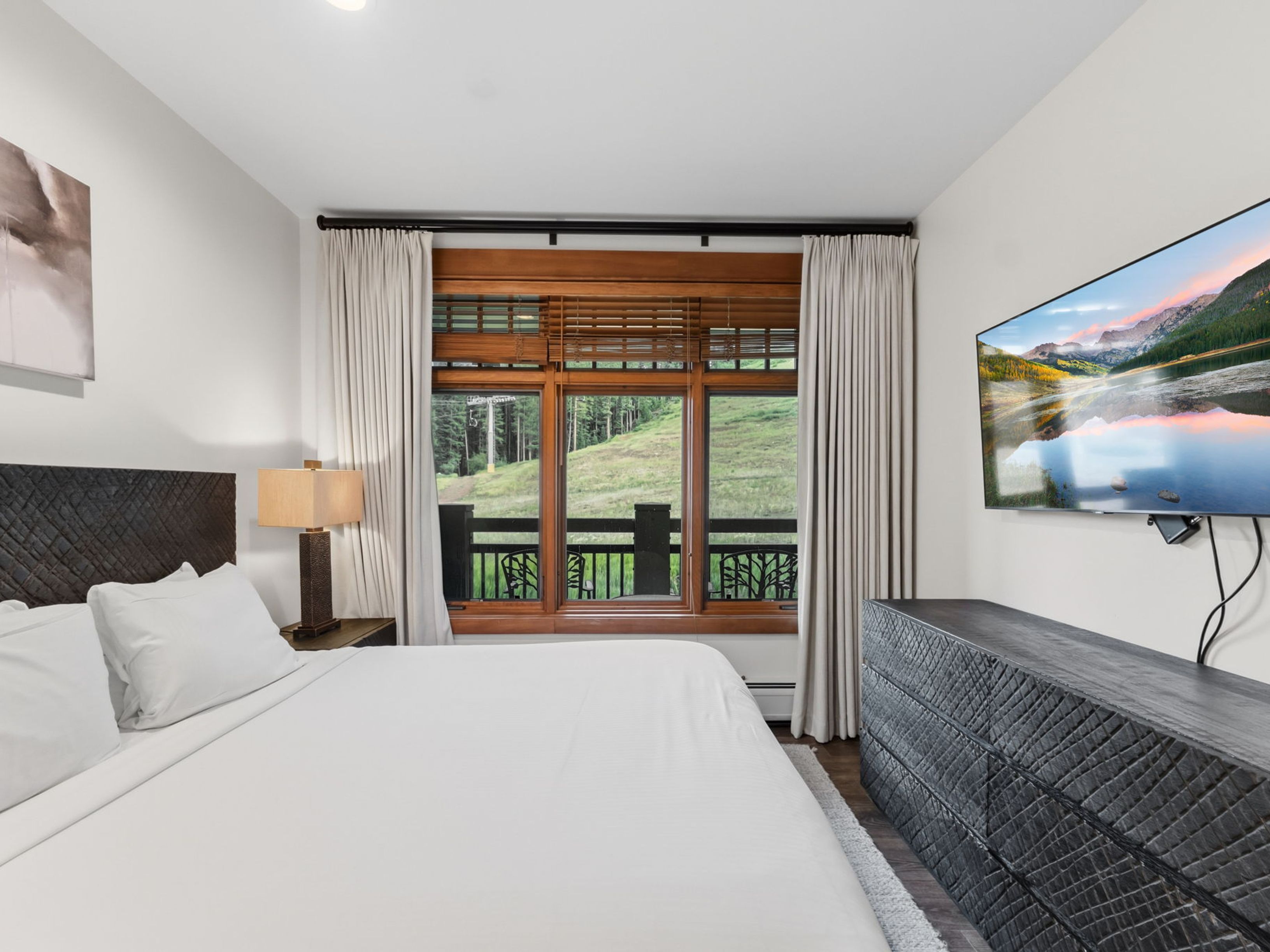 This primary bedroom features a large bed with a dark, textured headboard and crisp white linens, positioned centrally in the room. A dark, matching textured dresser sits beneath a wall-mounted television, while a bedside lamp provides warm lighting. Large windows framed by neutral curtains offer a scenic view of a grassy hillside and ski lift, creating a serene and inviting atmosphere.