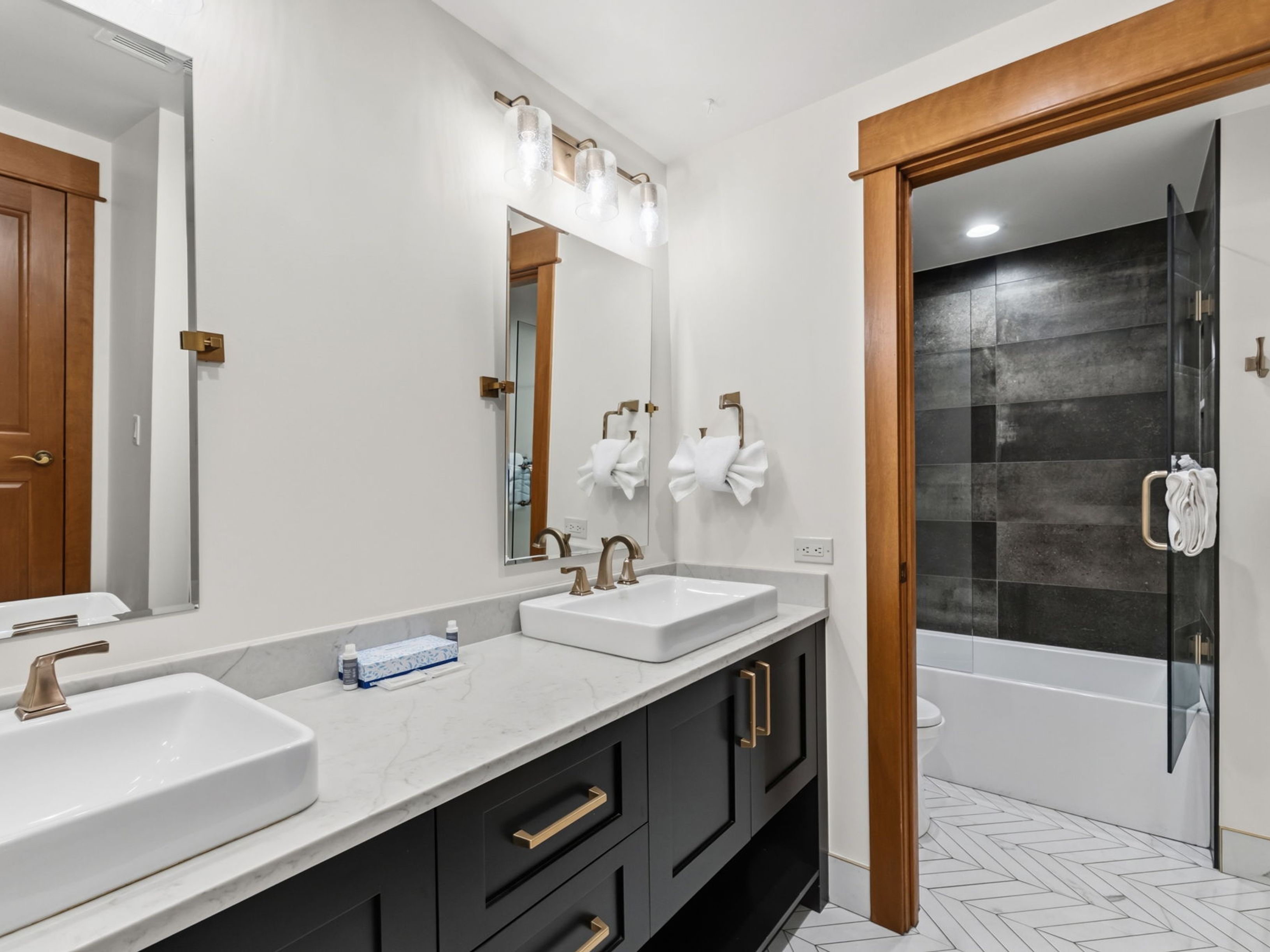This modern bathroom features a double vanity with dark cabinetry, gold-toned hardware, and a light-colored stone countertop with dual vessel sinks. The space is illuminated by a contemporary three-light fixture above the mirrors and includes a doorway leading to a separate shower area with dark, horizontal-tiled walls and a herringbone-patterned floor. The overall aesthetic is clean and sophisticated, blending warm wood trim with high-contrast finishes.