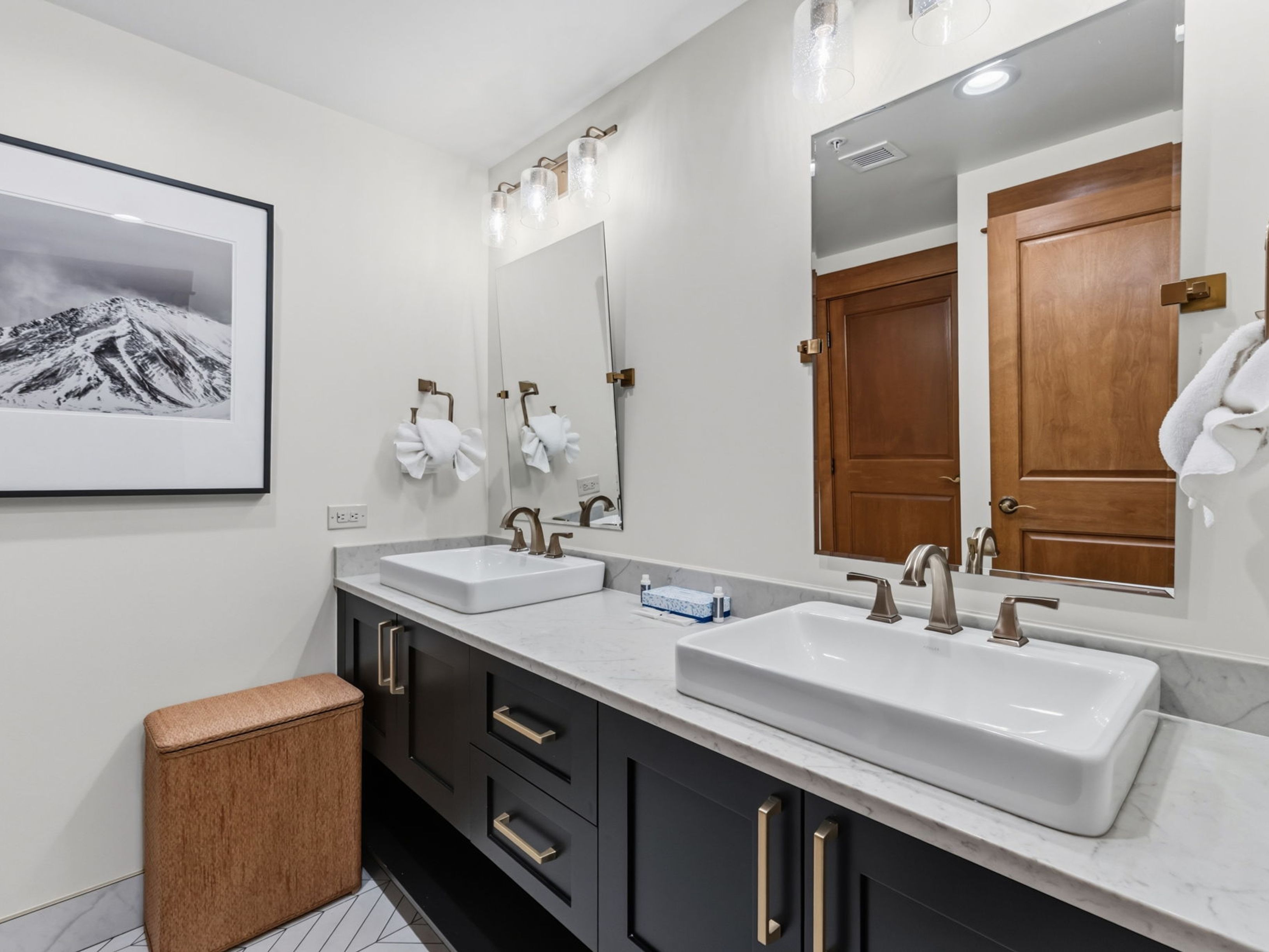 This modern double-vanity bathroom features dark cabinetry contrasted by a light stone countertop and two rectangular vessel sinks. The space is accented with warm-toned metal hardware, including faucets and drawer pulls, and includes a framed mountain landscape print on the wall. A large mirror reflects the wooden door and the clean, contemporary aesthetic of the room.