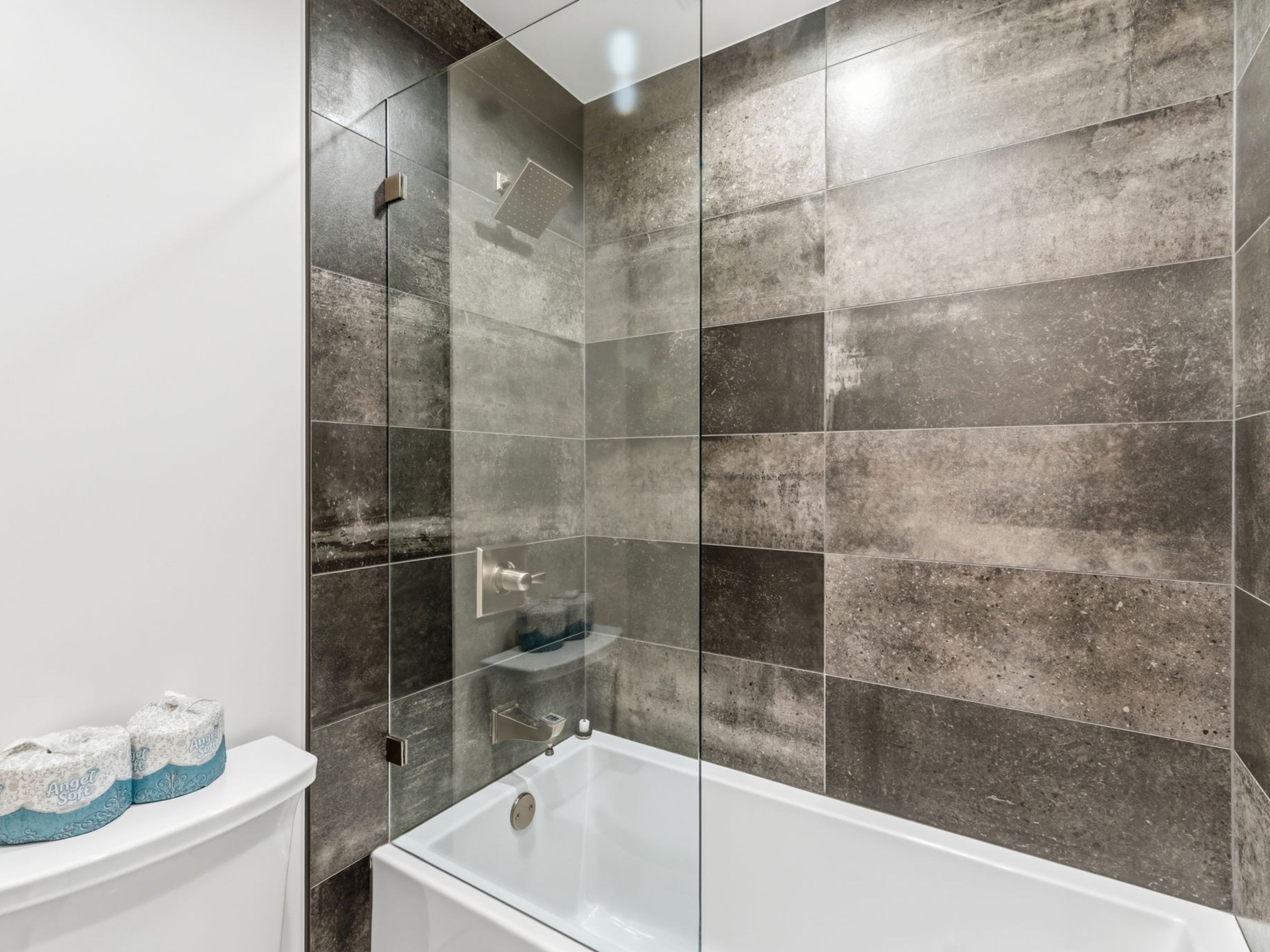 This bathroom features a modern shower-tub combination enclosed by a frameless glass panel. The walls are finished with large, rectangular tiles in varying shades of gray and charcoal, creating a textured, industrial-inspired aesthetic. A white toilet is positioned in the foreground, with two rolls of toilet paper resting on the tank, providing a clean and functional perspective of the space.