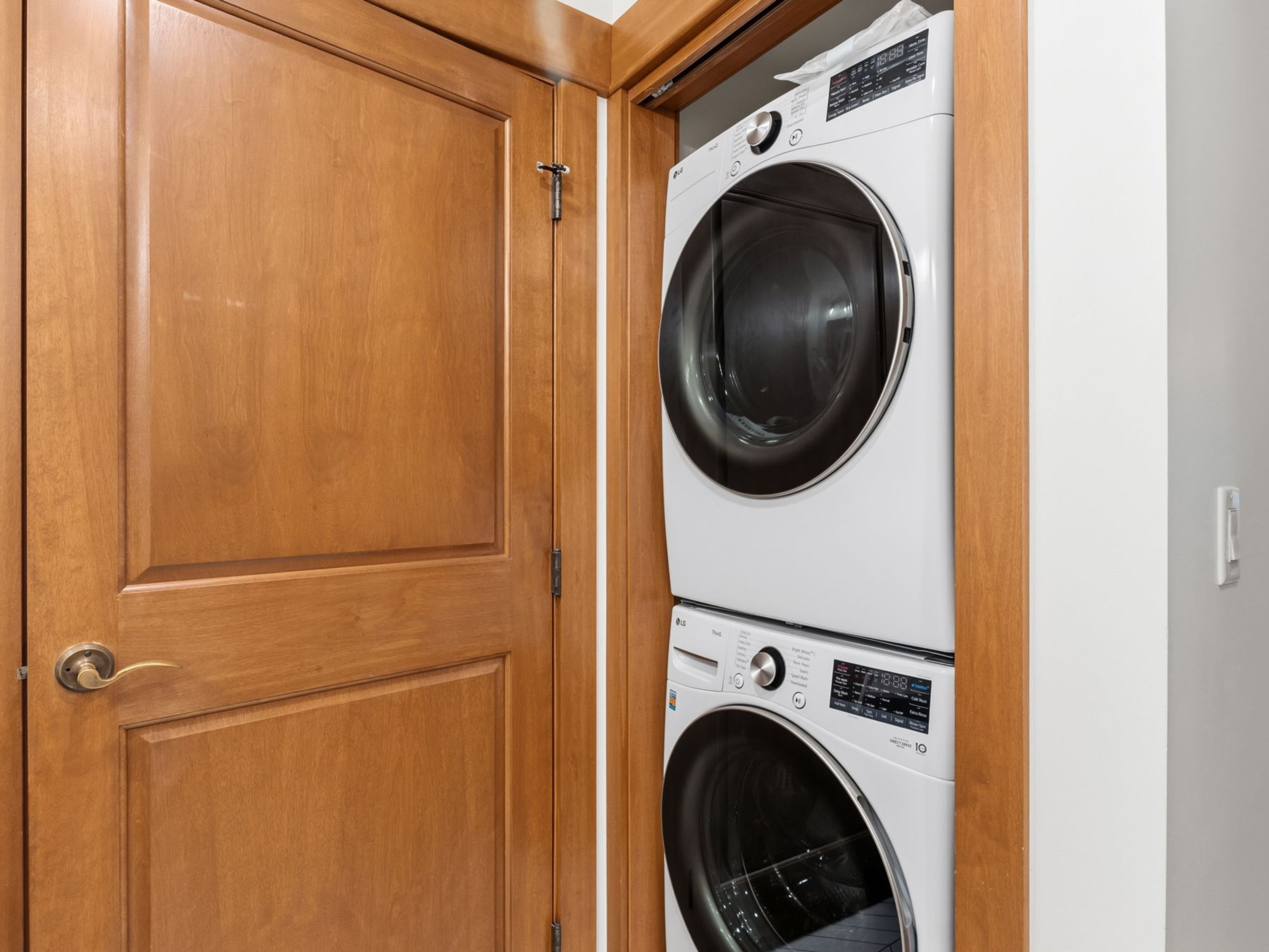 This image features a stacked washer and dryer unit neatly tucked into a dedicated closet space. The appliances are white with dark circular doors, set against a light-colored wall and framed by warm-toned wood trim. To the left, a matching wood door with a brass handle adds a cohesive, traditional touch to the utility area.
