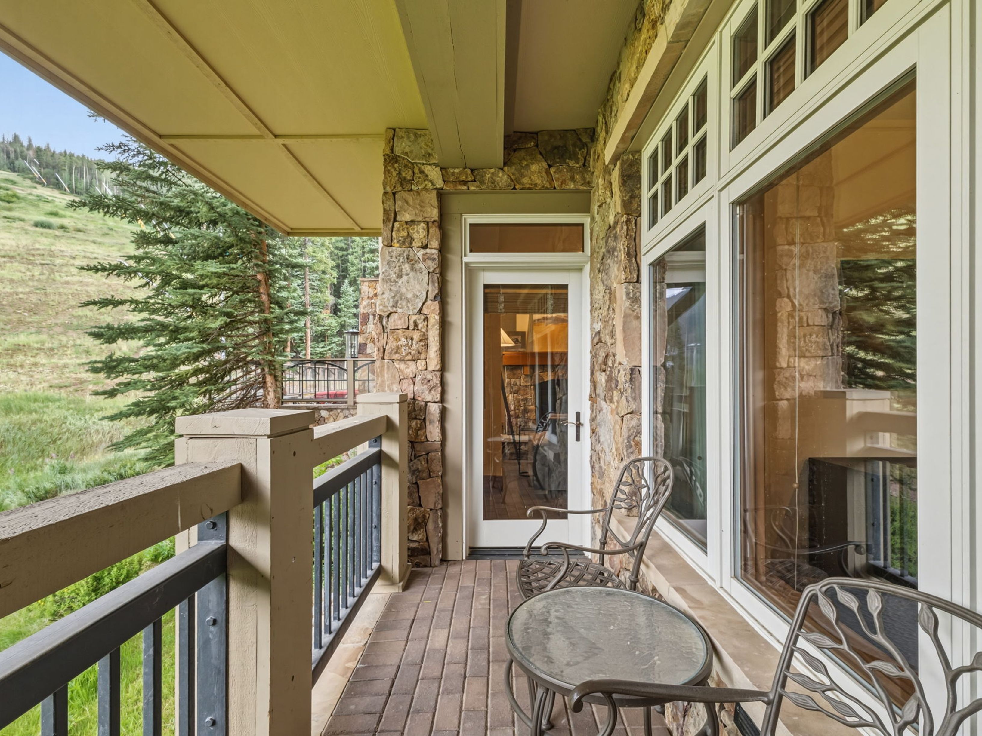 This covered balcony offers a serene outdoor living space with a view of a grassy ski slope and surrounding pine trees. The area features a brick-tiled floor, a stone-clad wall, and a metal railing, complemented by a small bistro set for relaxing. Large windows and a glass-paneled door provide a seamless transition to the interior, creating an inviting atmosphere for mountain living.