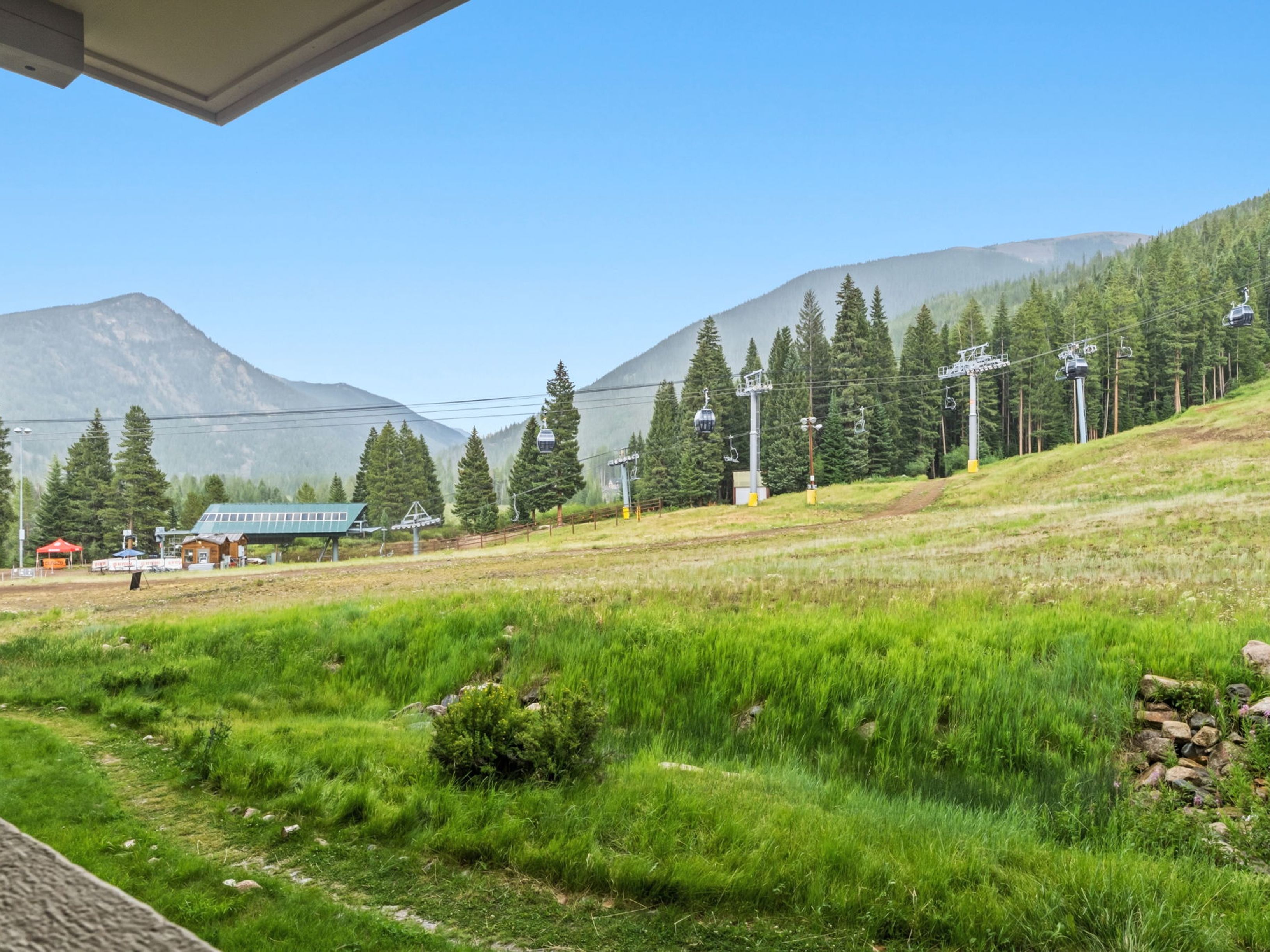This view from an elevated balcony captures a scenic mountain landscape featuring a ski lift system and lush green slopes. The foreground shows a grassy meadow leading toward the base of the ski area, where a lift station and small structures are visible. The perspective provides a wide, open look at the recreational amenities and natural surroundings, suggesting a prime location for outdoor enthusiasts.