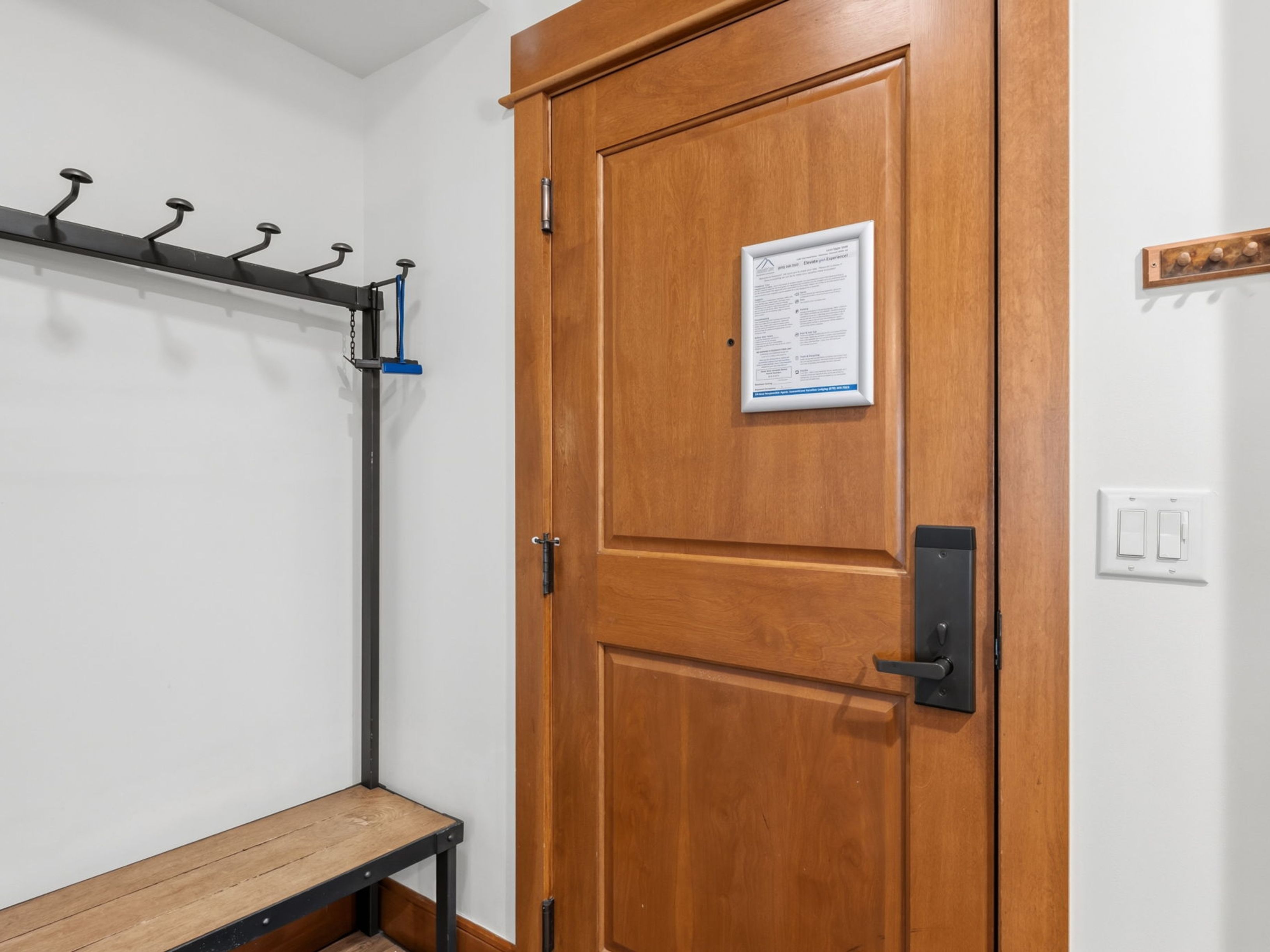 This entryway features a warm-toned wooden door equipped with a modern black lever handle and a posted informational sign. To the left, a functional mudroom area includes a black metal coat rack with multiple hooks mounted above a simple wooden bench. The space is finished with clean white walls, a light switch panel, and a small decorative wooden rack on the right wall, creating a practical and welcoming transition area.