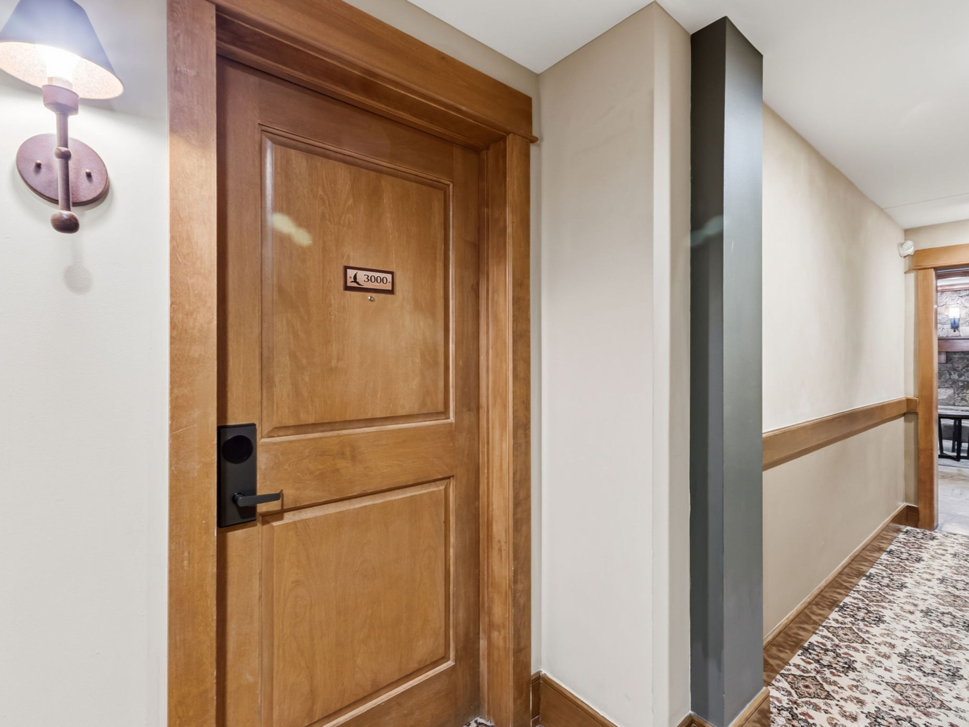This image shows a hallway featuring a wooden door marked with the number 3000, accented by a wall-mounted light fixture. The hallway includes a dark-painted pillar and a patterned carpet runner, leading toward another doorway in the distance. The overall aesthetic is traditional and well-maintained, typical of a hotel or residential corridor.