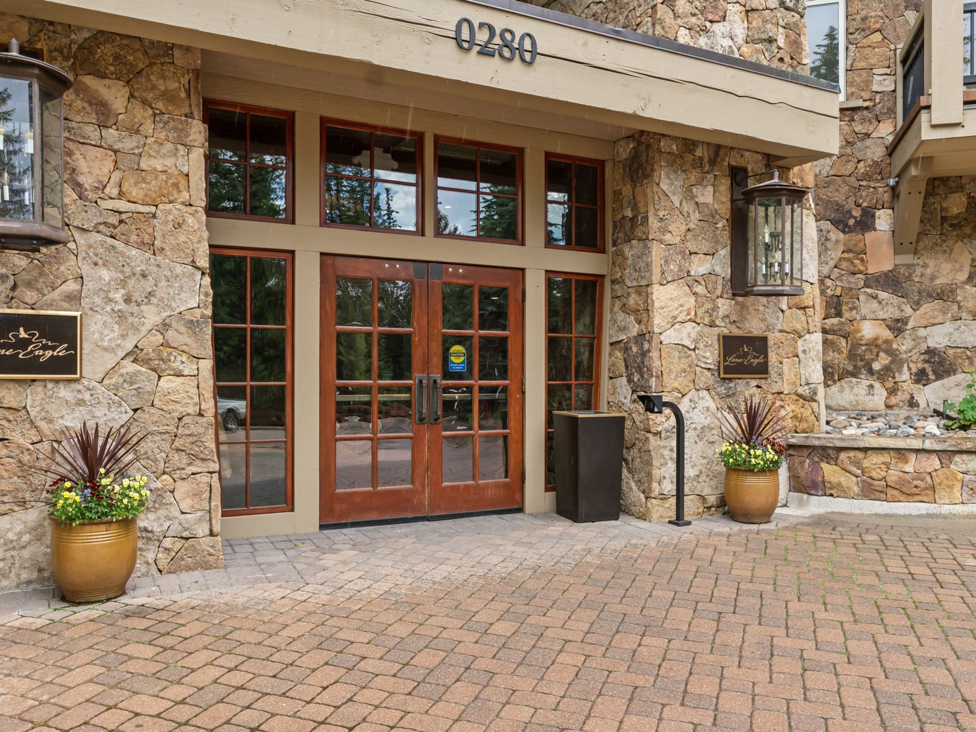 This inviting entryway features a rustic stone facade with a prominent wooden double-door entrance marked by the address '0280'. The exterior is accented by large, dark-framed windows, decorative wall-mounted lanterns, and two large planters filled with vibrant flowers flanking the doorway. The ground is paved with a textured stone paver driveway, creating a warm and welcoming arrival experience.