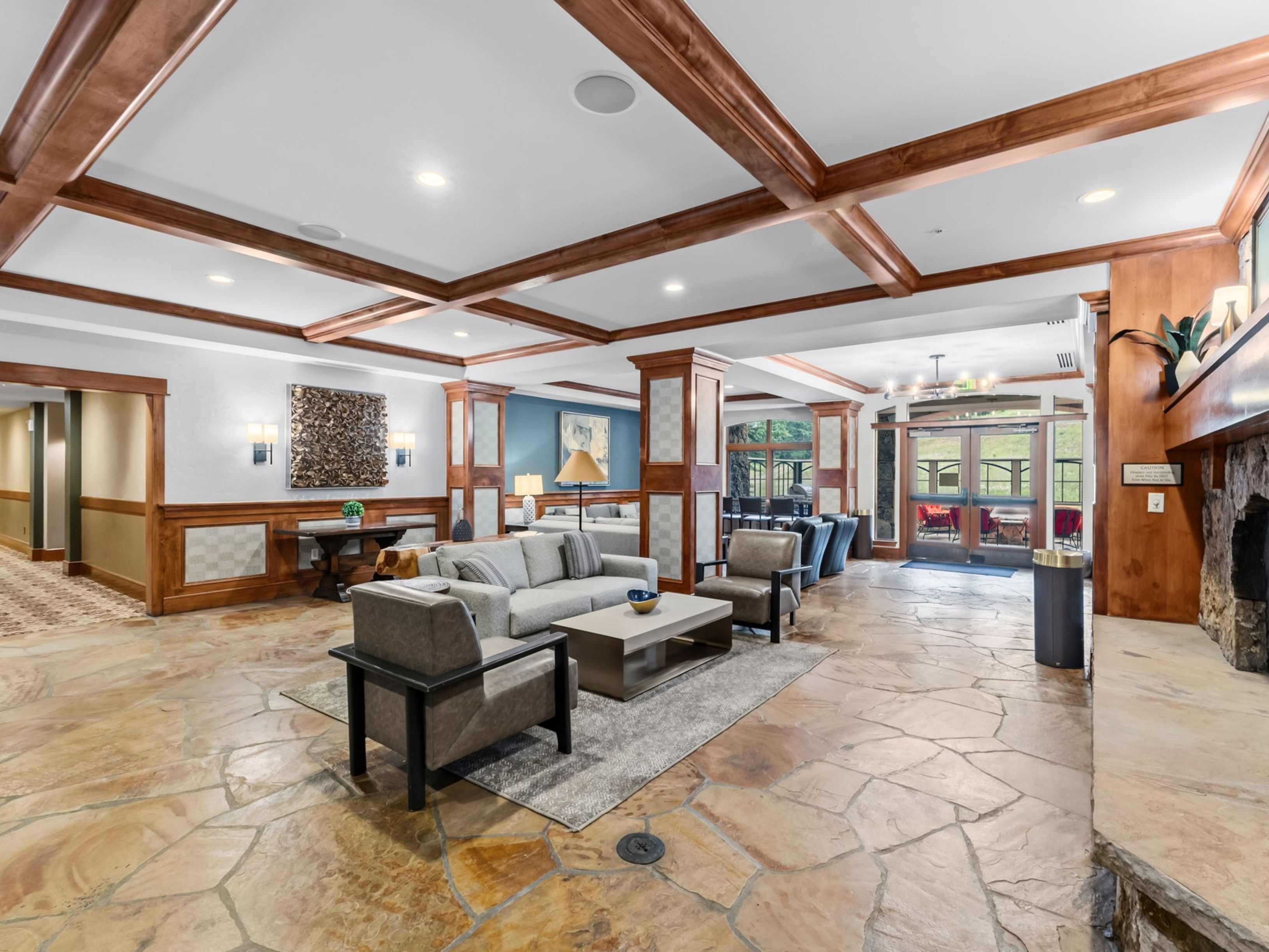 This spacious lobby or clubhouse lounge features a rustic aesthetic with a stone-tiled floor and a coffered ceiling accented by dark wood beams. The seating area is furnished with a neutral-toned sofa and armchairs arranged around a low coffee table, while a stone-clad chimney breast stands to the right. Large glass doors at the rear provide a view of the outdoor landscape, creating an inviting and open atmosphere for residents or guests.