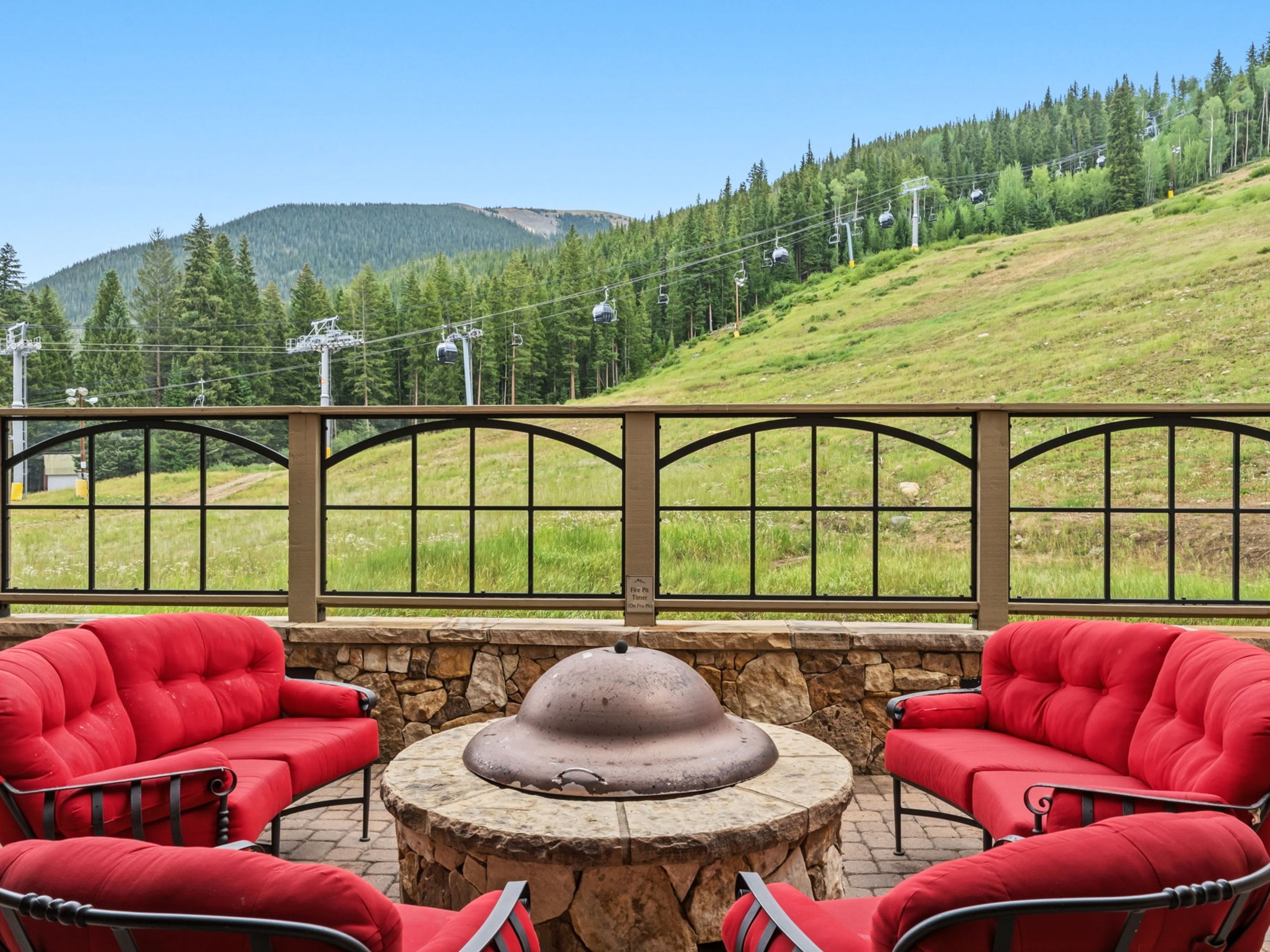 This outdoor patio space features a stone-clad fire pit as the central focal point, surrounded by four vibrant red cushioned sofas. The area is enclosed by a decorative metal railing that overlooks a scenic mountain landscape with ski lift cables visible in the distance. The setting offers a relaxing, resort-style atmosphere perfect for enjoying the expansive views of the grassy hillside.