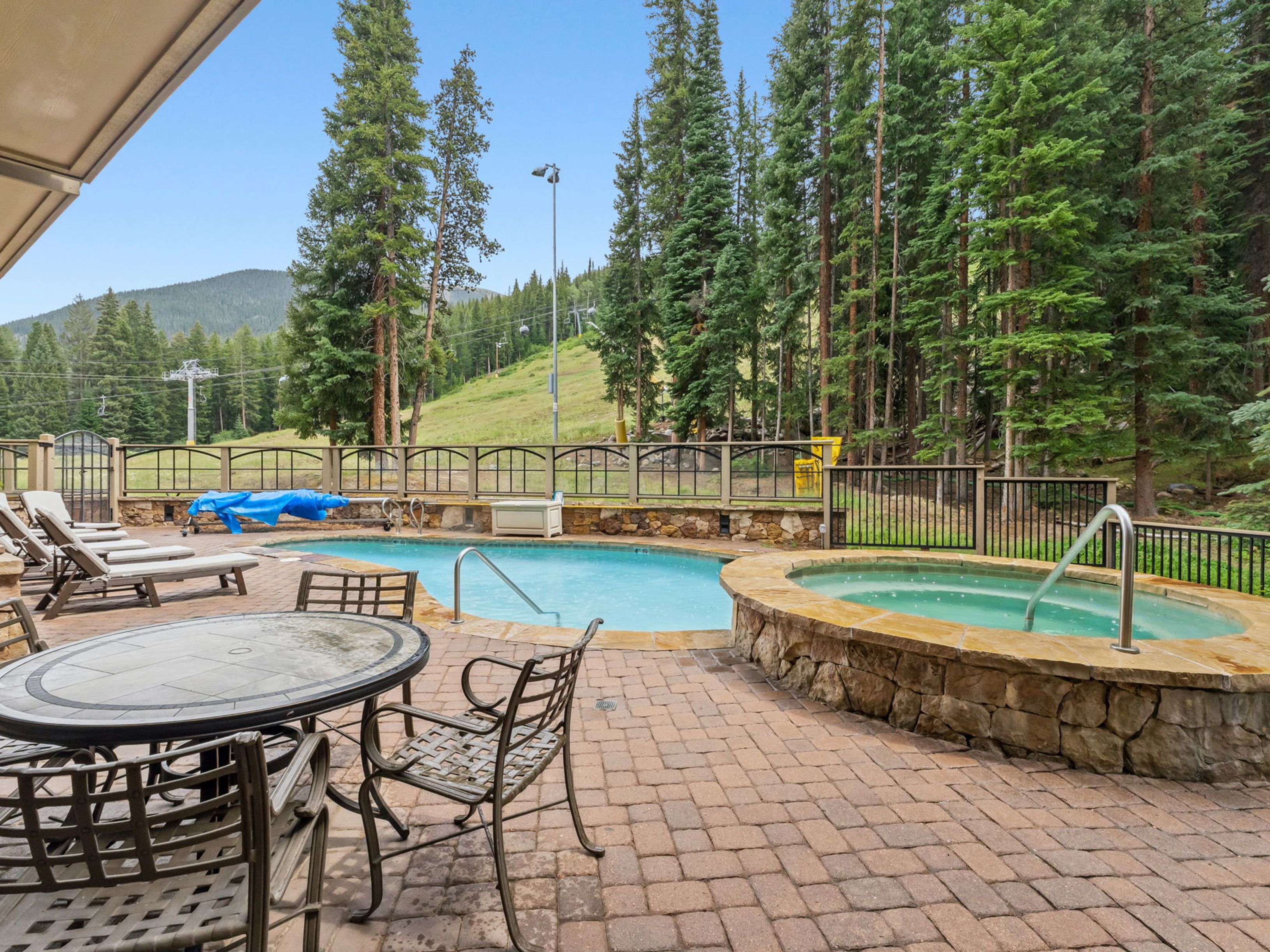 This outdoor patio area features a swimming pool and a circular stone-clad hot tub, set against a backdrop of dense pine trees and a mountain ski slope. The space is furnished with a metal dining set and several lounge chairs, all arranged on a paved stone deck. The scene captures a serene, resort-like atmosphere perfect for relaxation with a clear view of the surrounding natural landscape.