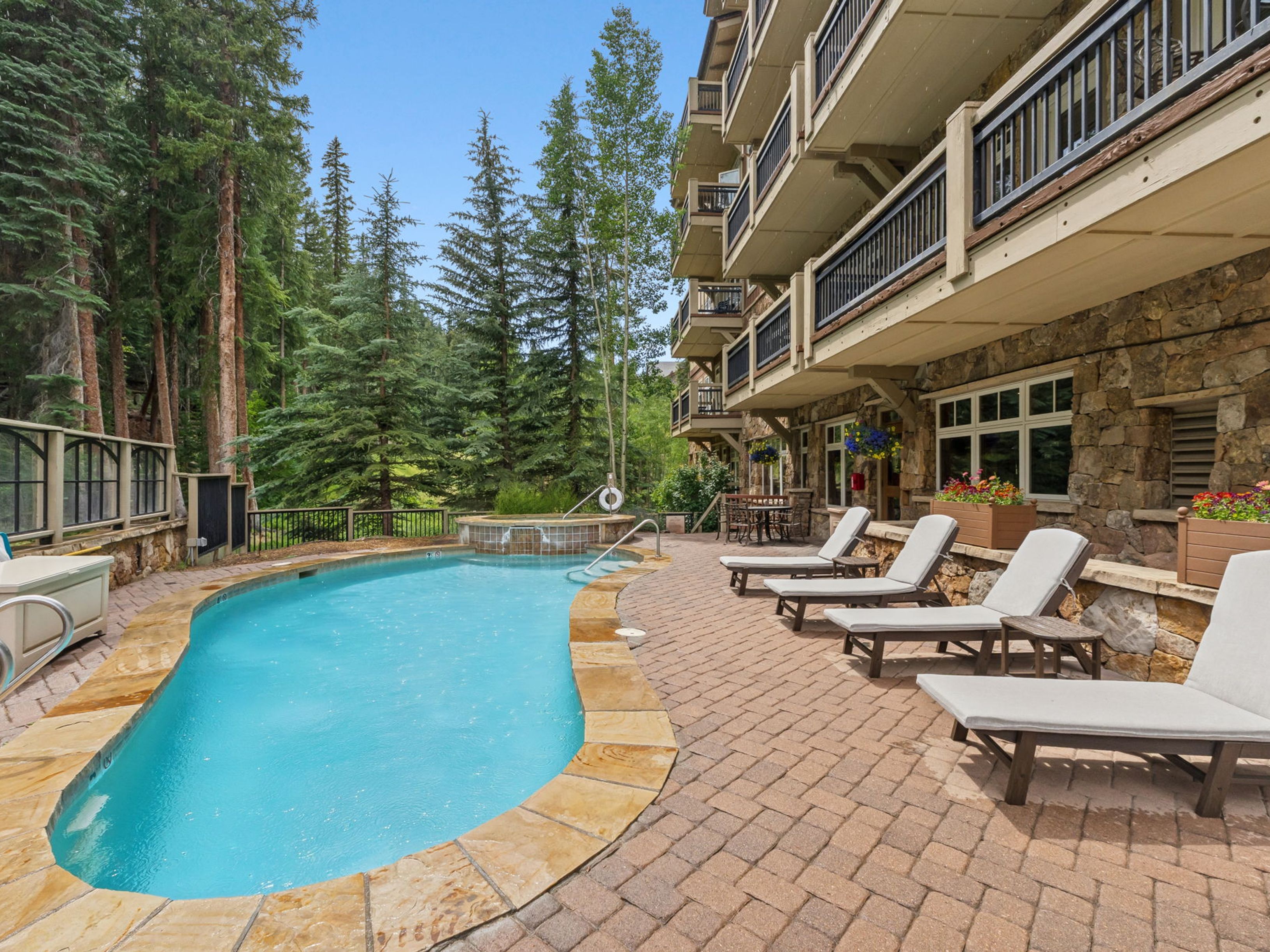 This outdoor amenity area features a curved swimming pool and an adjacent hot tub set against a multi-story building with stone-clad walls and dark metal railings. A paved patio area is furnished with several lounge chairs, offering a relaxing space for residents to enjoy the surrounding forest views. The scene is captured from a ground-level perspective, emphasizing the integration of the pool deck with the natural landscape.